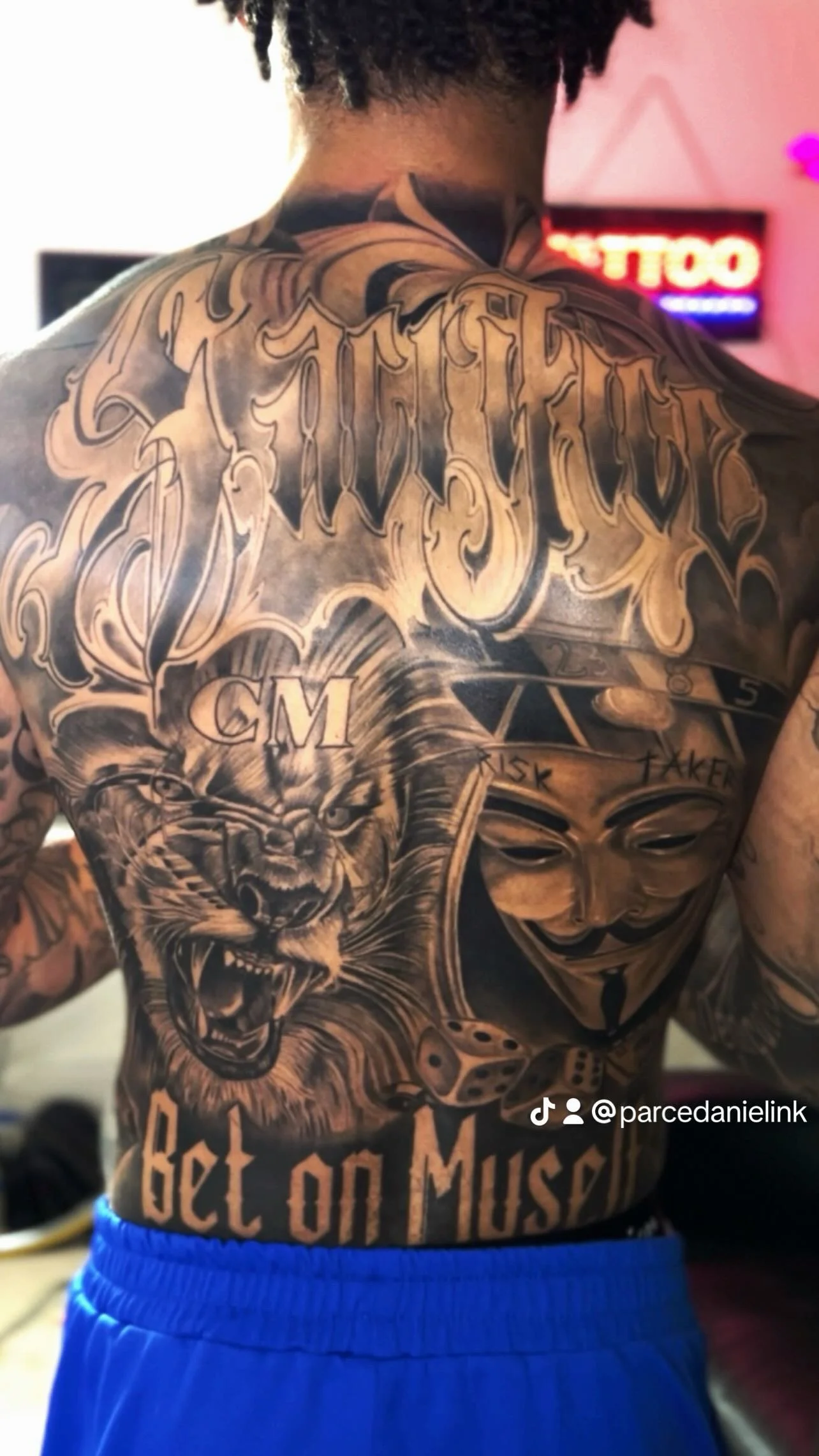 Close-up of a person's back and torso with extensive tattoos, including a lion's face, a girl wearing a Guy Fawkes mask, a lion, and various text and symbols. The person is wearing blue shorts.