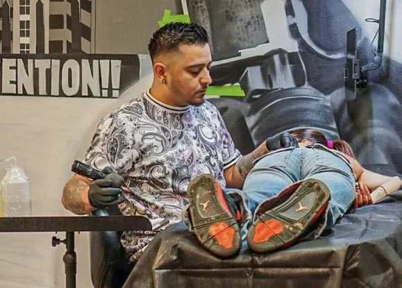Tattooed man in a patterned shirt giving a tattoo to a person lying on a tattoo table.