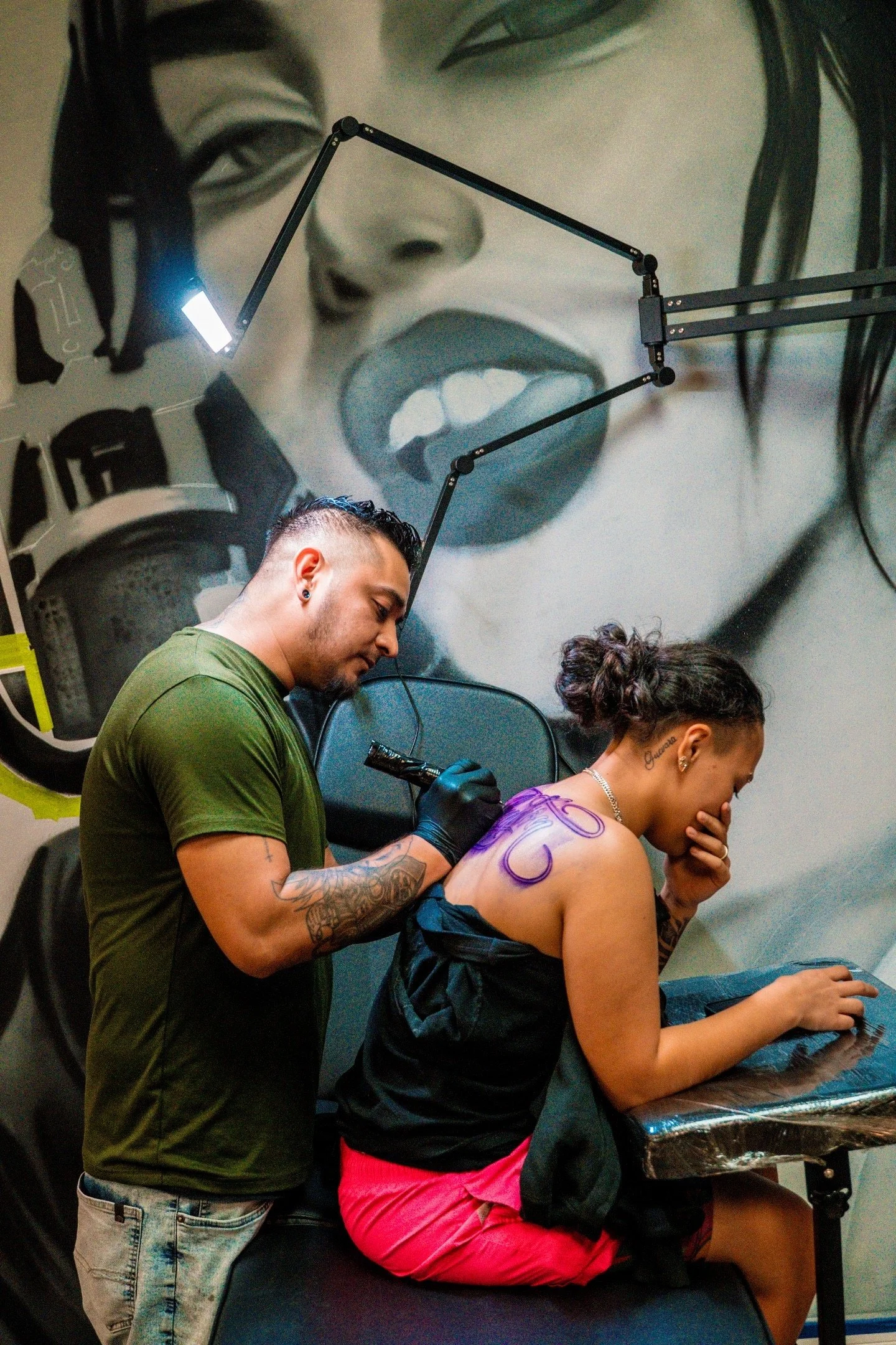 A person with tattoos on their left arm is tattooing the back of a woman who is seated and waiting. The woman has purple markings on her back and is looking down with a hand covering her mouth. A man is standing beside her, holding a tattoo gun, and 