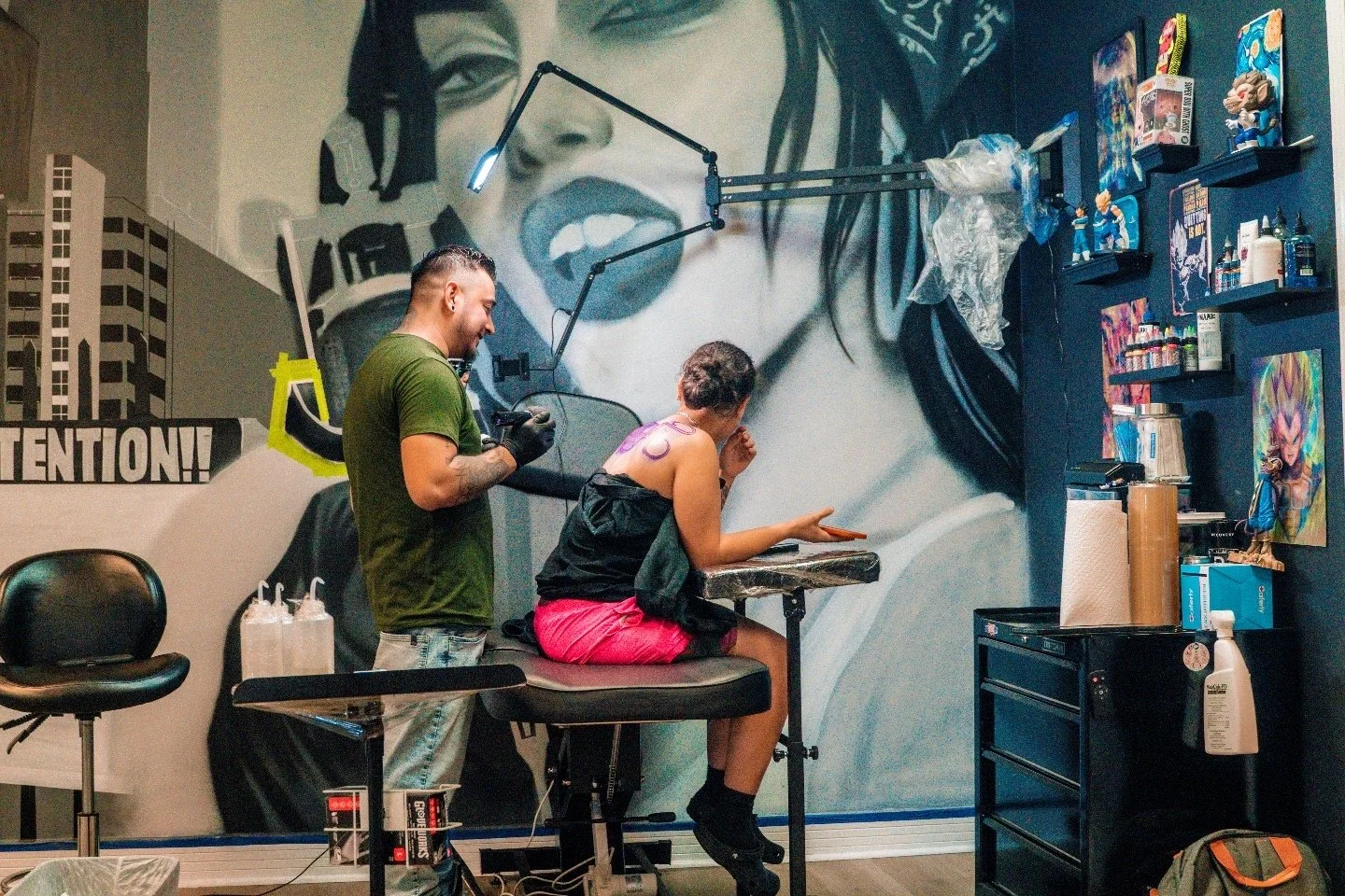 A woman getting tattooed by a tattoo artist in a studio. The woman is seated, holding a phone and appears to be relaxing or talking. The artist is standing, smiling, and preparing for the tattoo work. The studio has a large mural of a woman singing o