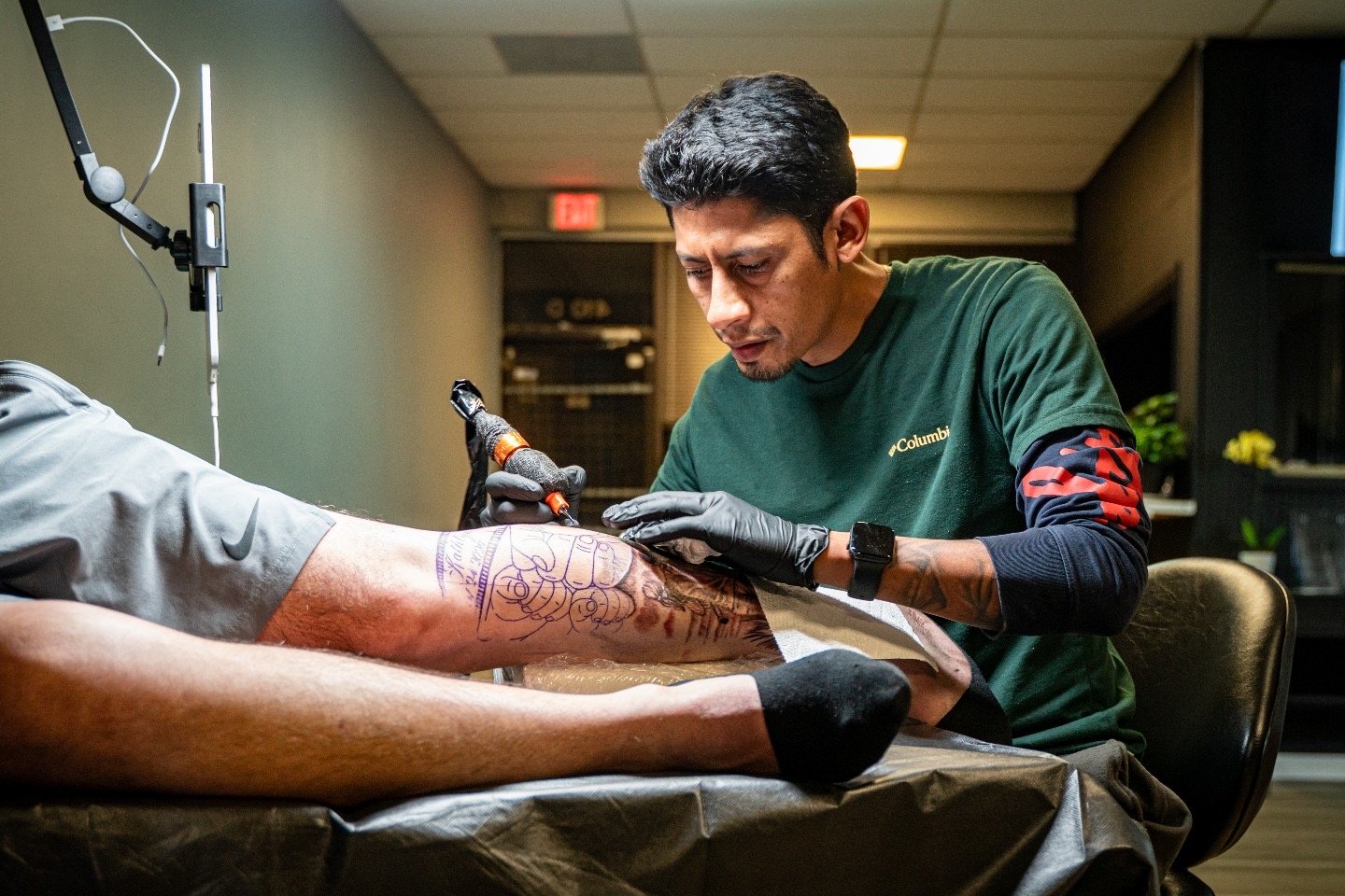 A tattoo artist working on a client's arm in a tattoo studio. The artist is focused, wearing black gloves and using a tattoo machine, with tattoo designs visible on the client's arm.