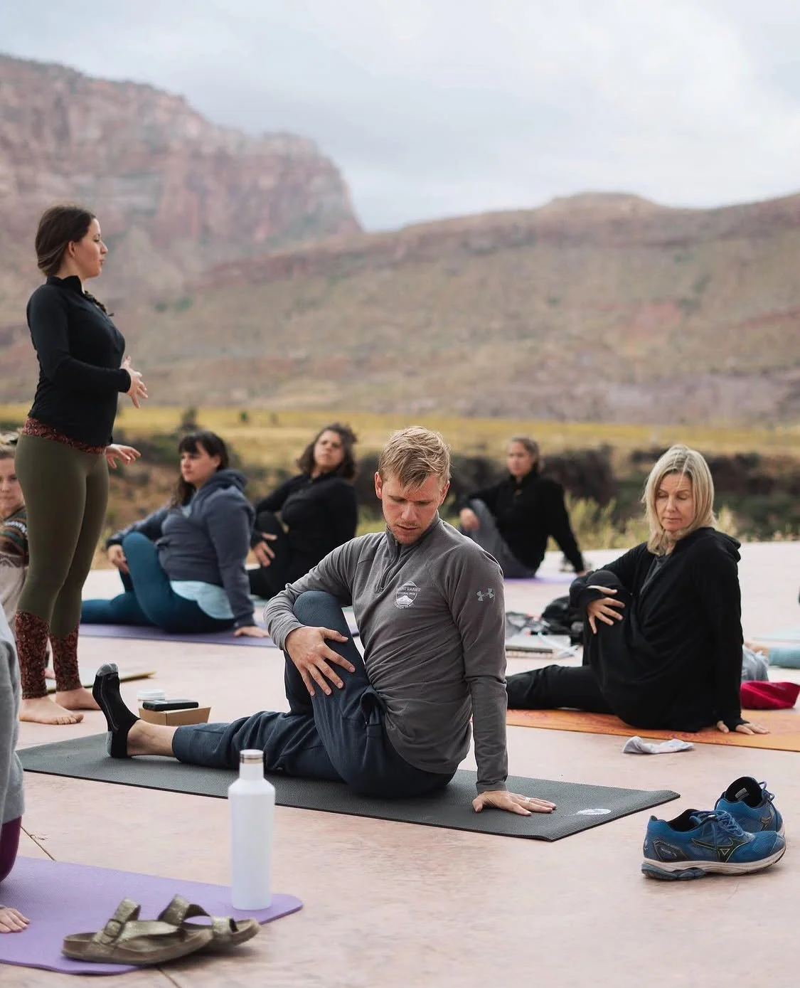 People participating in an outdoor yoga class on a scenic mountain landscape, with some seated on yoga mats and a woman standing and instructing.