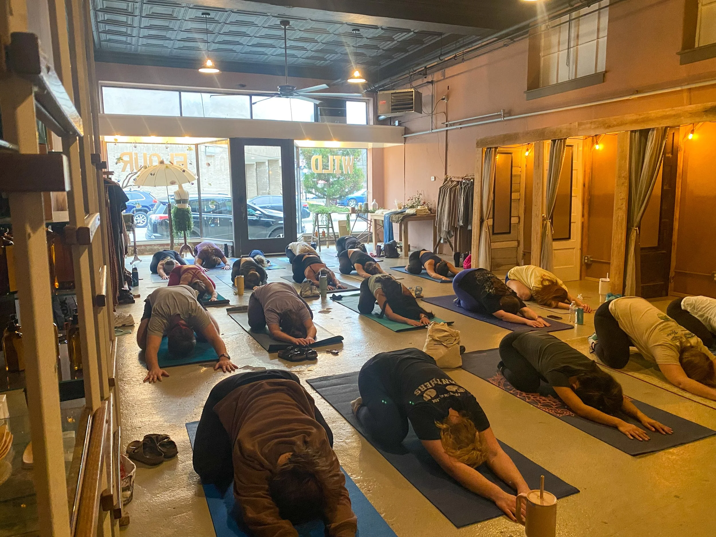 People participating in a yoga class or meditation session in a warmly lit indoor space with large windows, practicing prone pose on mats, with some clothing racks and decor in the room.