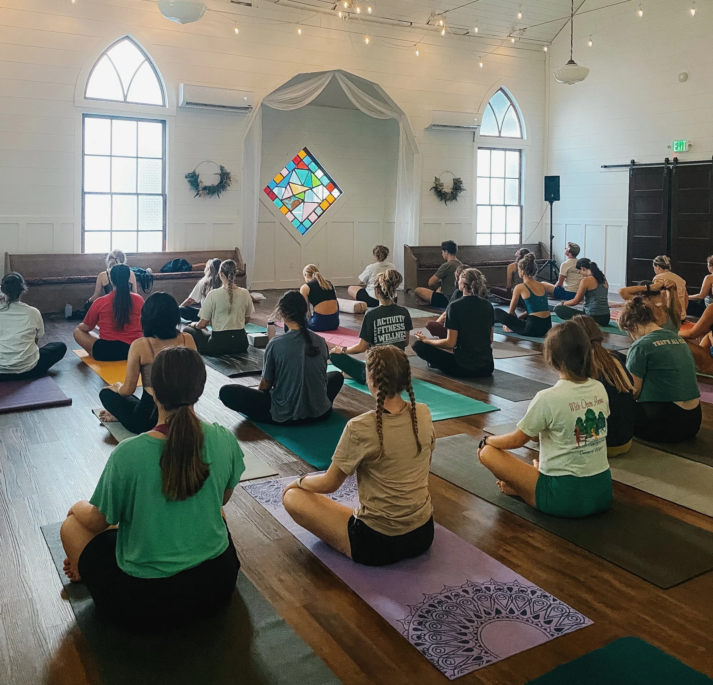 People seated on yoga mats inside a bright room with arched windows, string lights, and a stained glass window at the front, participating in a group meditation or yoga session.