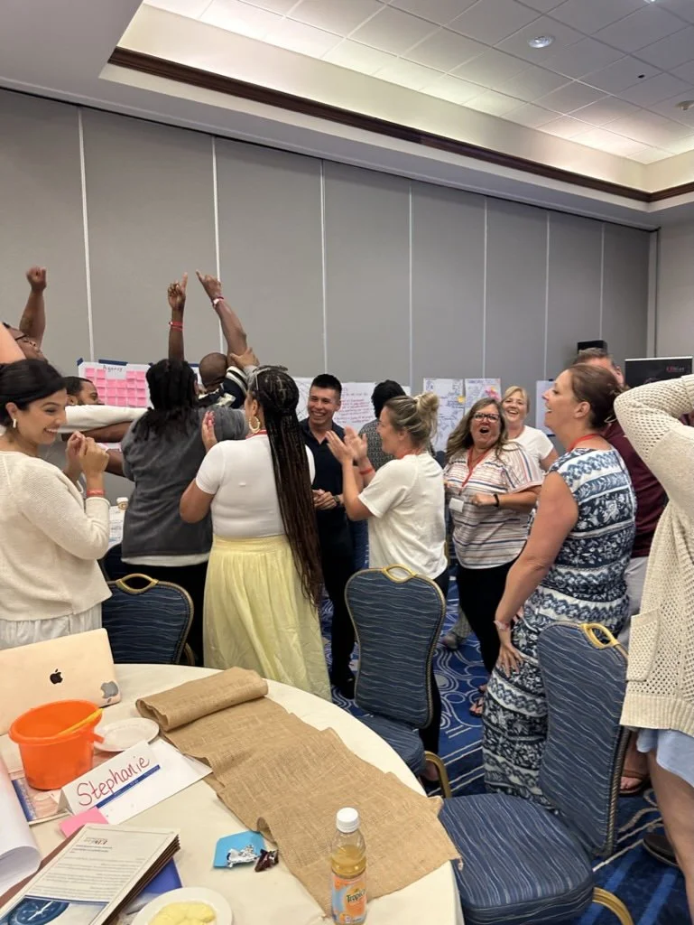 A photo of Erin Wills leading a workshop for educational leaders. The group is cheering and excited around post-it boards.