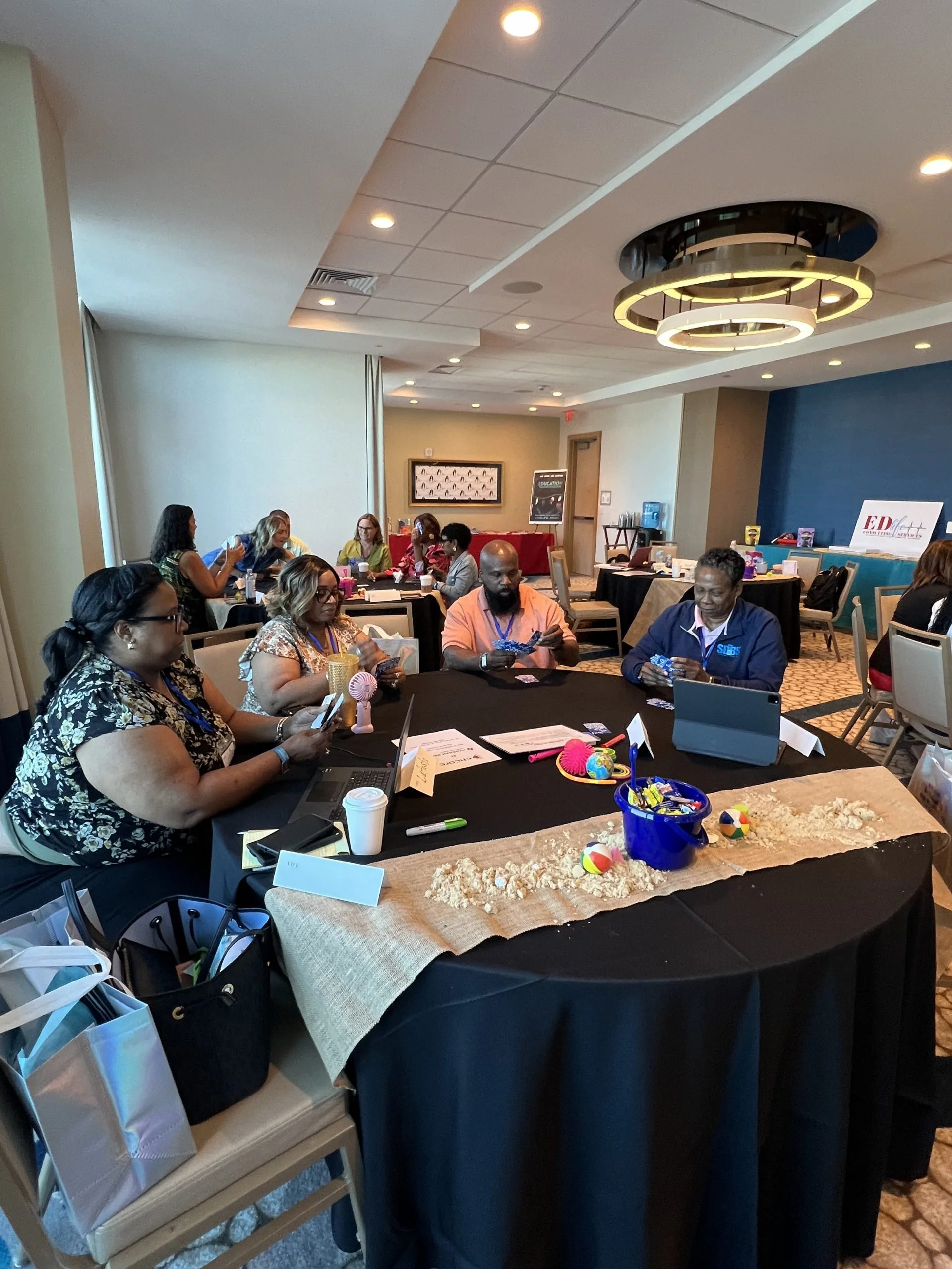 People gathered in a conference room playing a game with beach balls, toys, and snacks on a covered table.