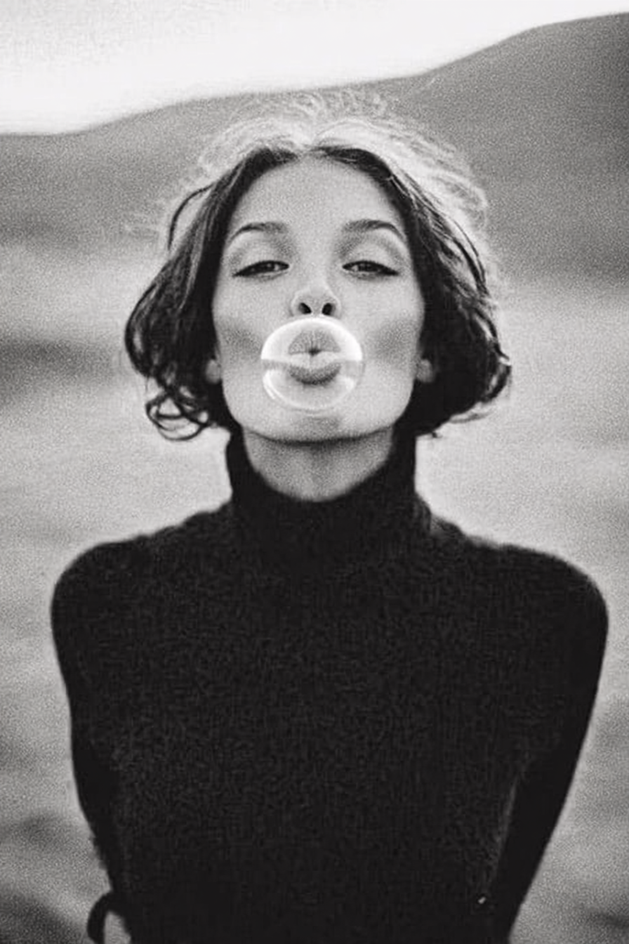 Black and white photo of a woman with short wavy hair blowing a bubble gum bubble, wearing a turtleneck sweater.