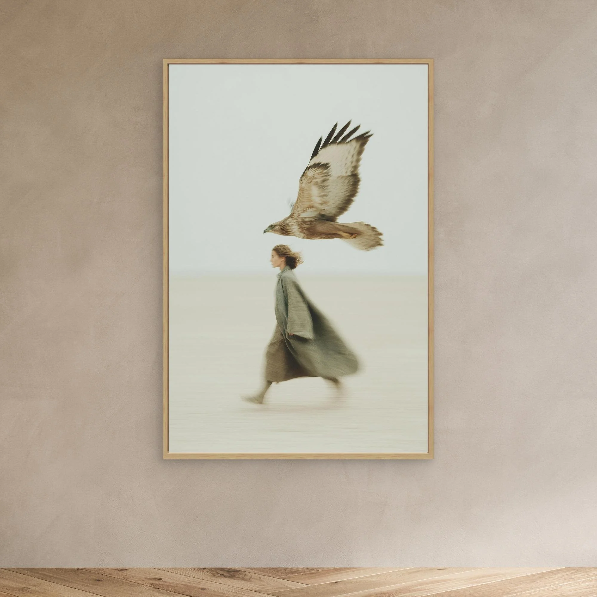 A framed artwork of a woman walking with an eagle flying above her, mounted on a beige wall in a room with wooden flooring.