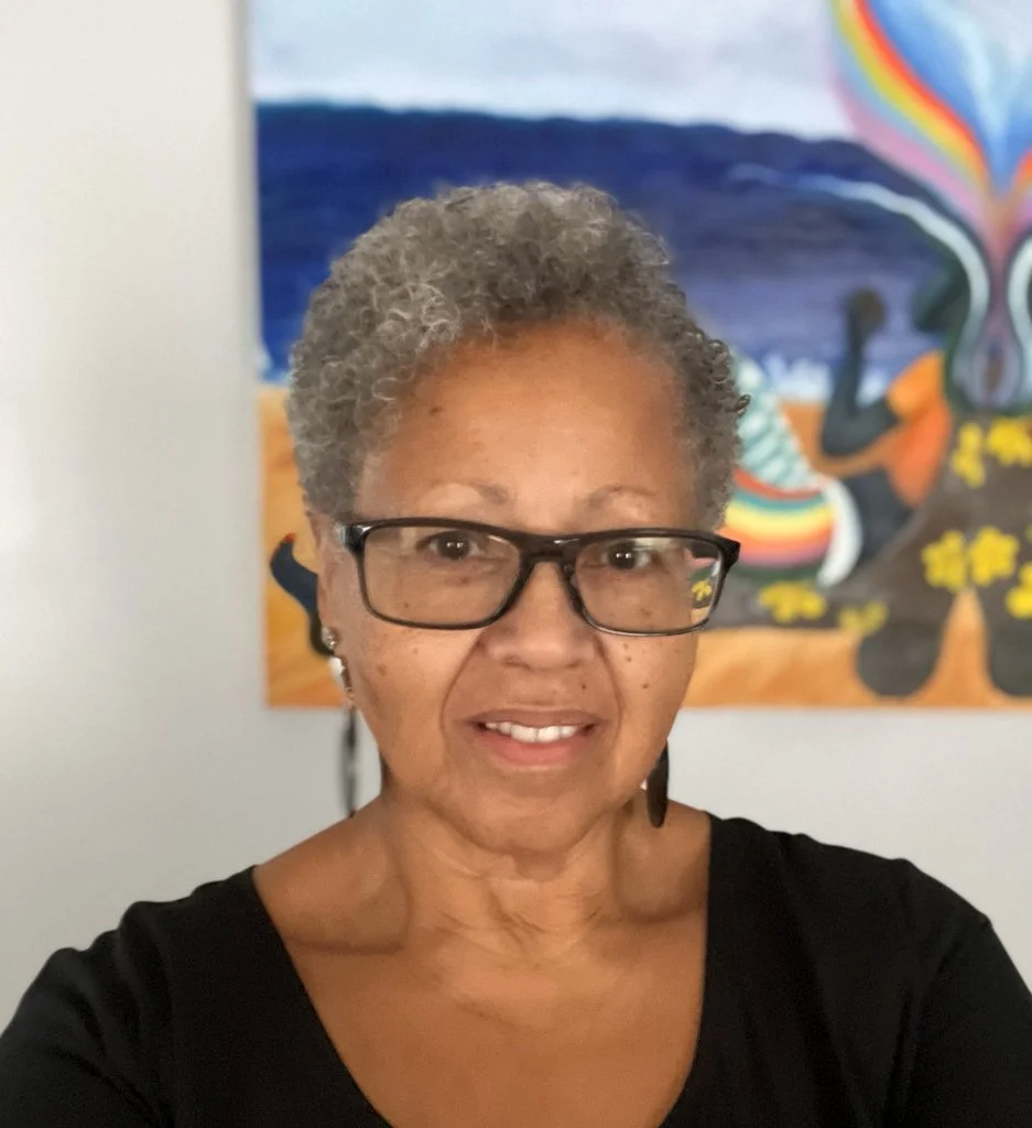A woman with short gray hair and glasses taking a selfie in front of a colorful painting of a beach scene with a rainbow, flowers, and a butterfly.