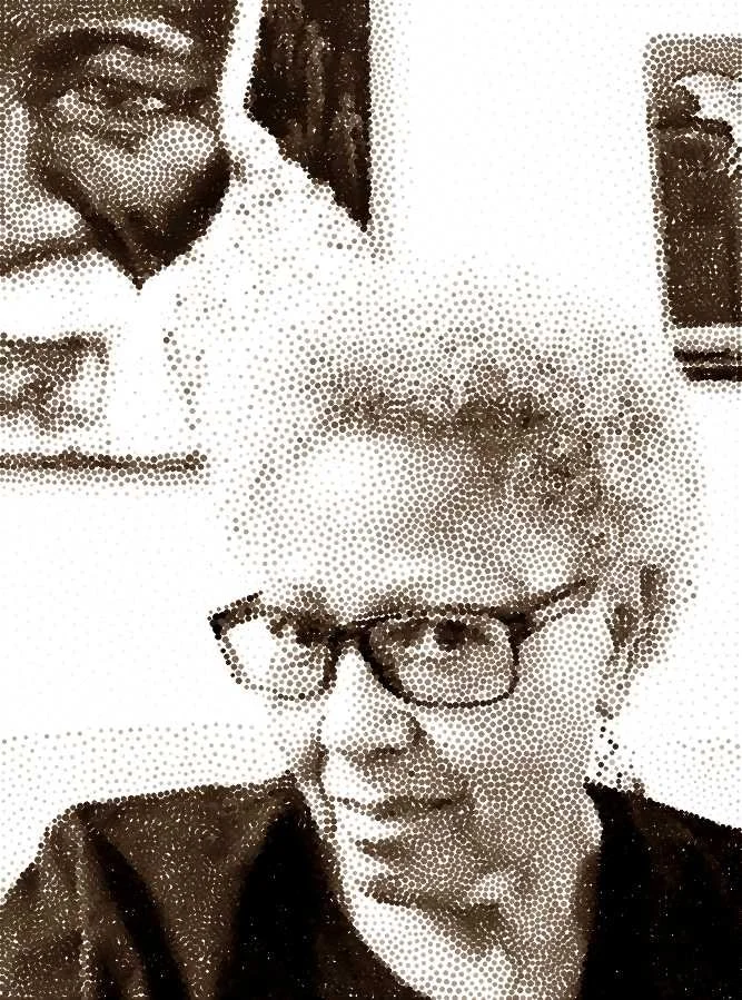 A woman with short blonde hair and glasses taking a selfie. There is a man smiling in the background.