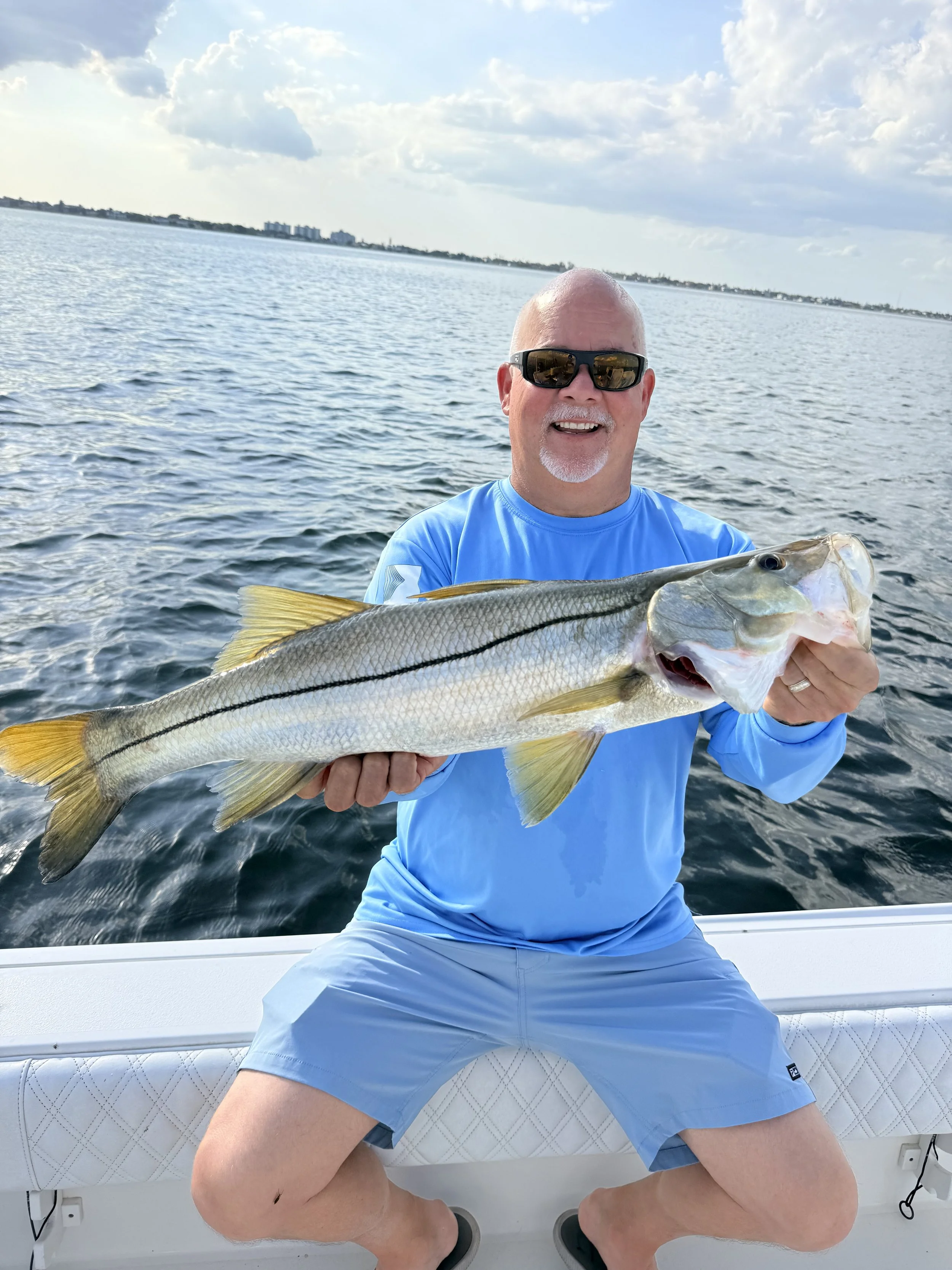A man with sunglasses holding a large fish onboard a boat on the water with a cloudy sky in the background.