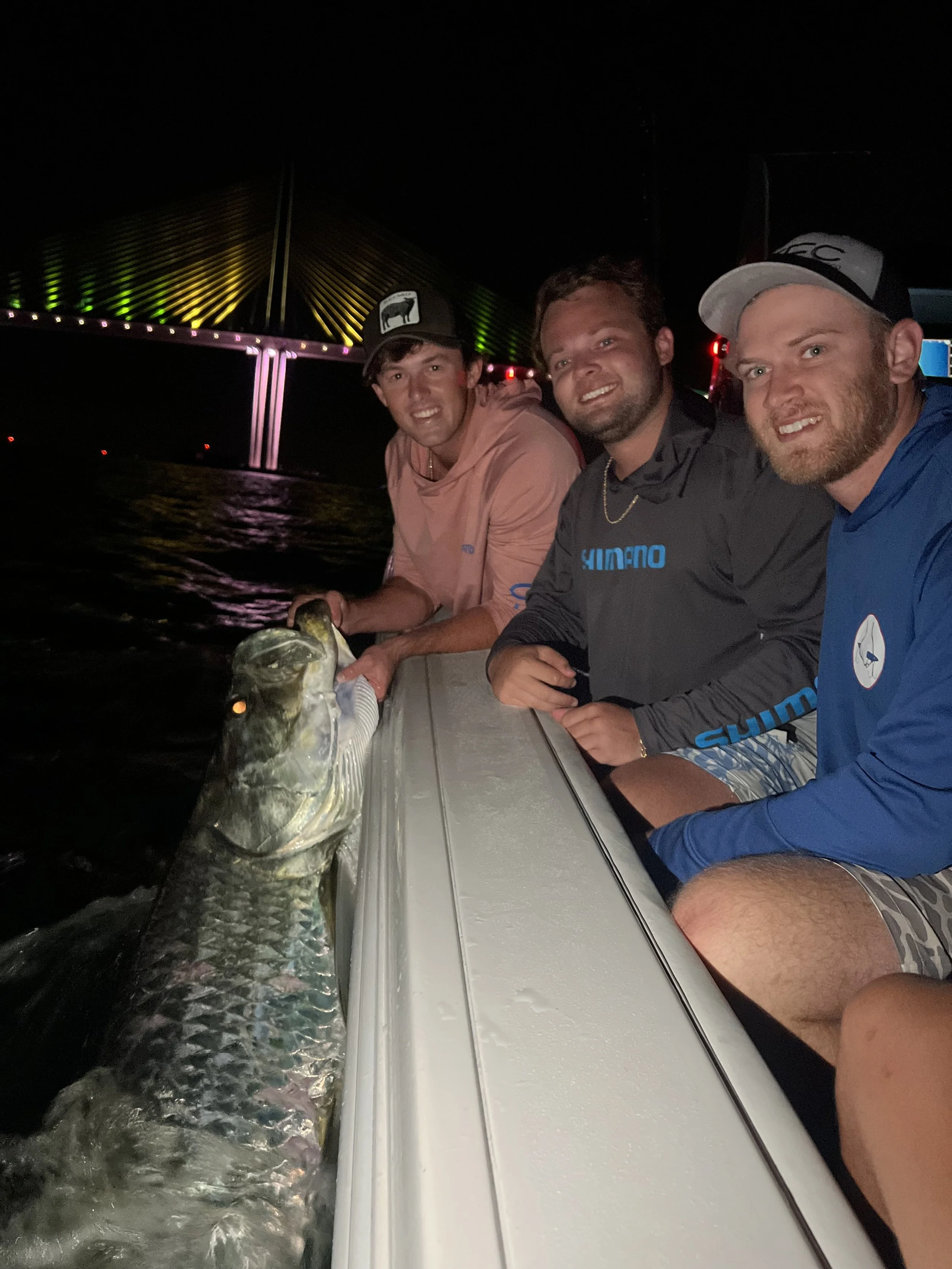 Three men sitting on a boat at night, holding a large fish they caught, with a brightly lit bridge in the background.