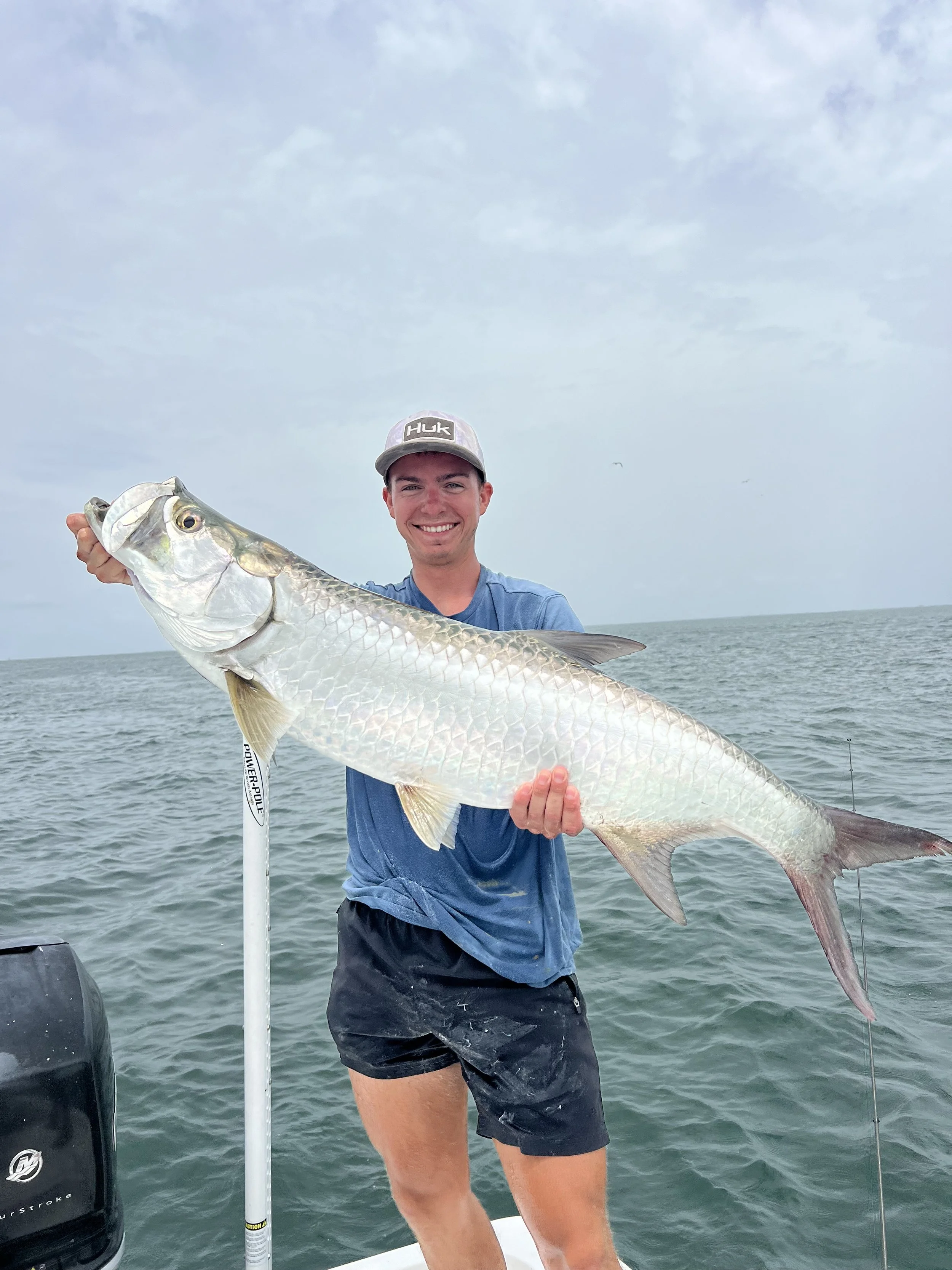 A man wearing a grey HUK cap, blue shirt, and black shorts, smiling and holding a large fish on a boat in the ocean during a cloudy day.