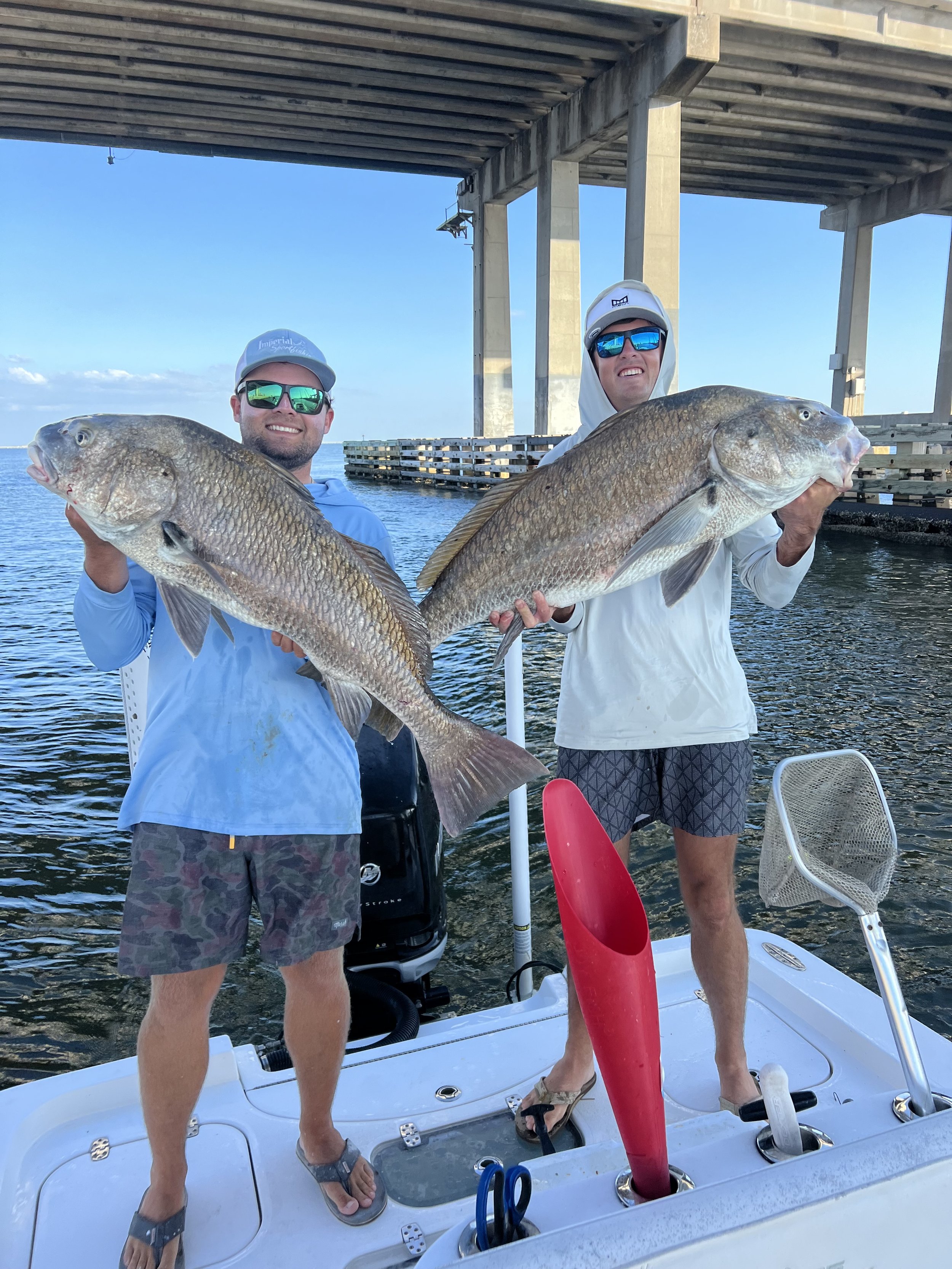 Two men on a boat holding large fish they caught, standing under a bridge with water in the background, wearing sunglasses and casual clothing.
