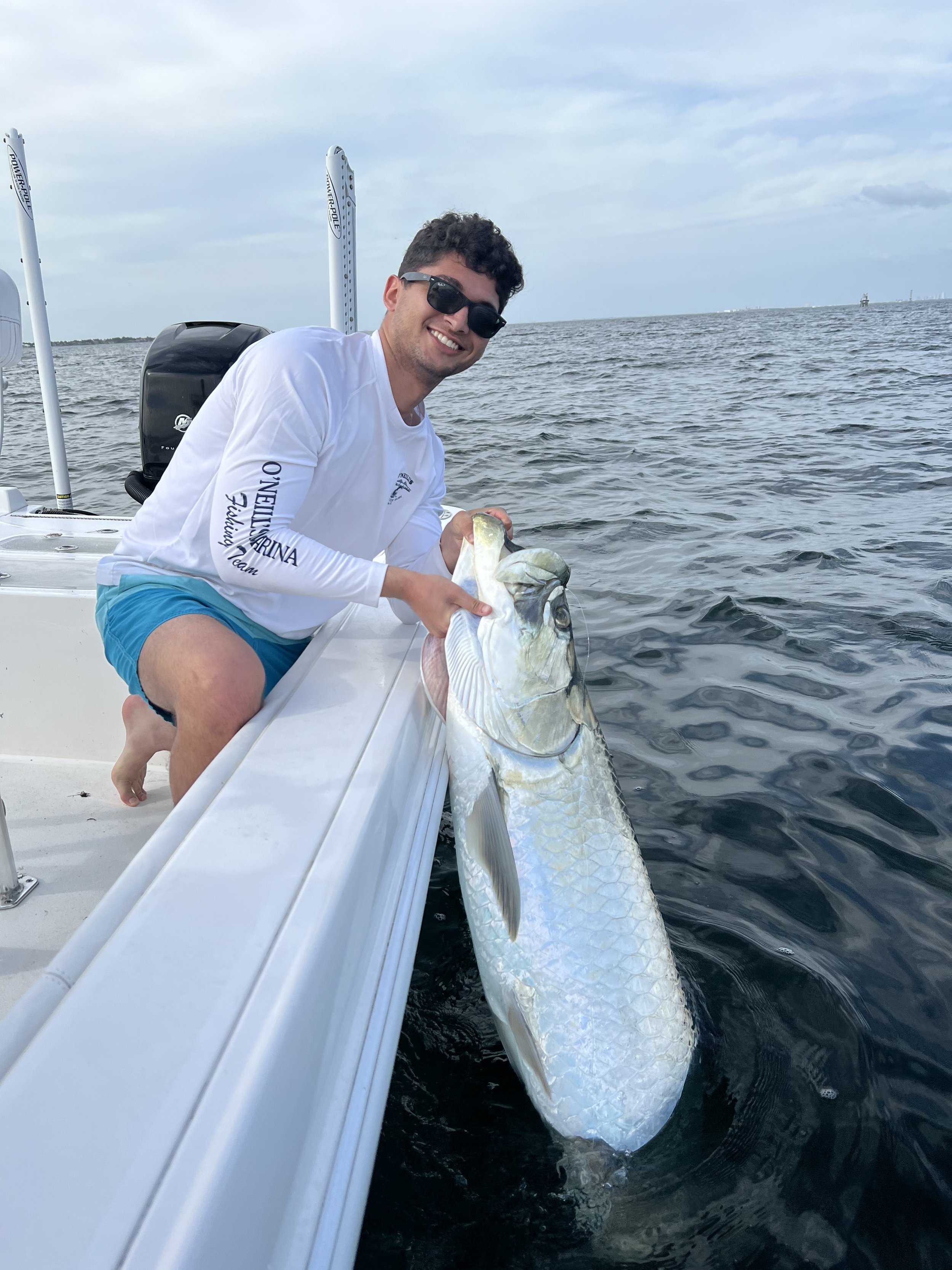 A smiling man wearing sunglasses, a white long-sleeve shirt, and blue shorts, sitting on a boat, holding a large fish he just caught, with the ocean and cloudy sky in the background.