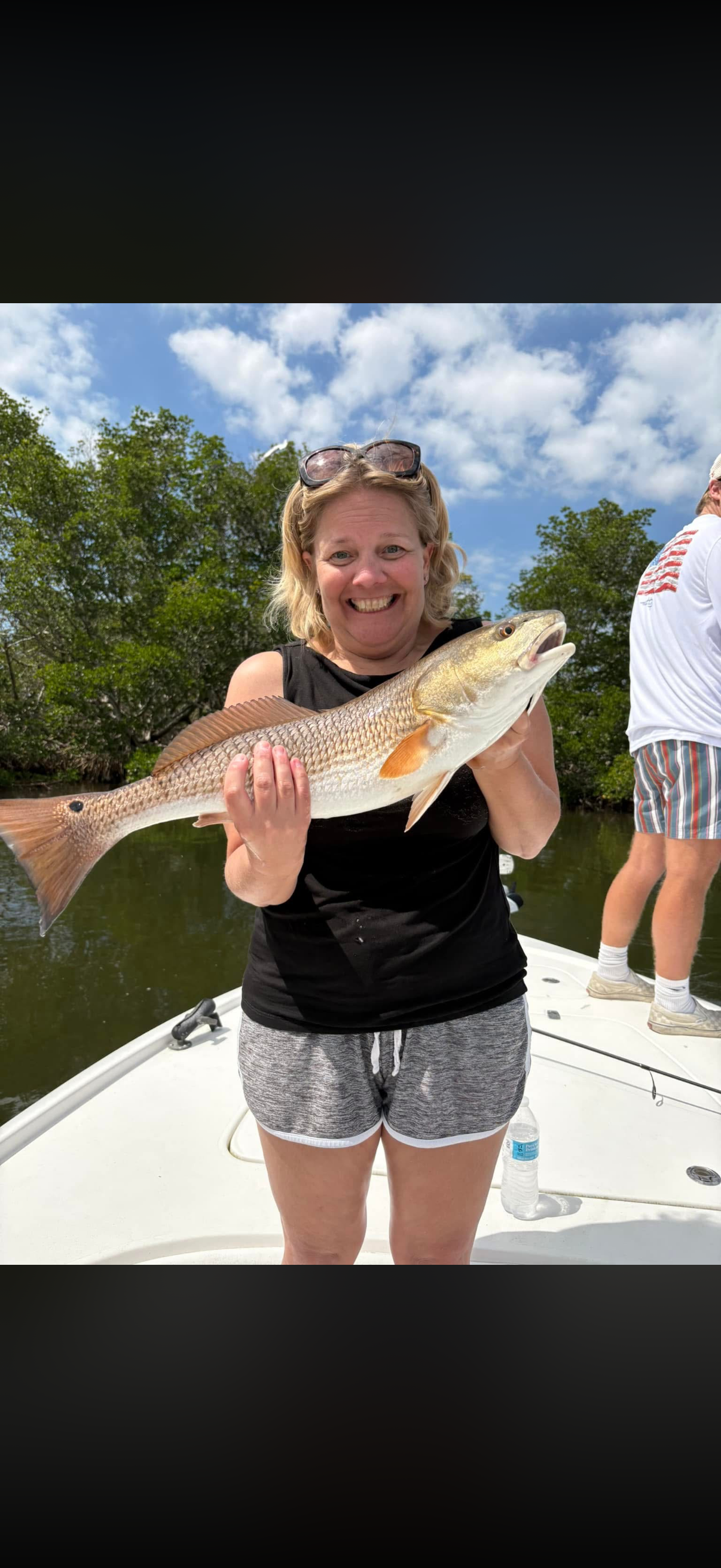 A smiling woman in a black tank top and gray shorts holding a large fish on a boat, with trees and a partly cloudy sky in the background.