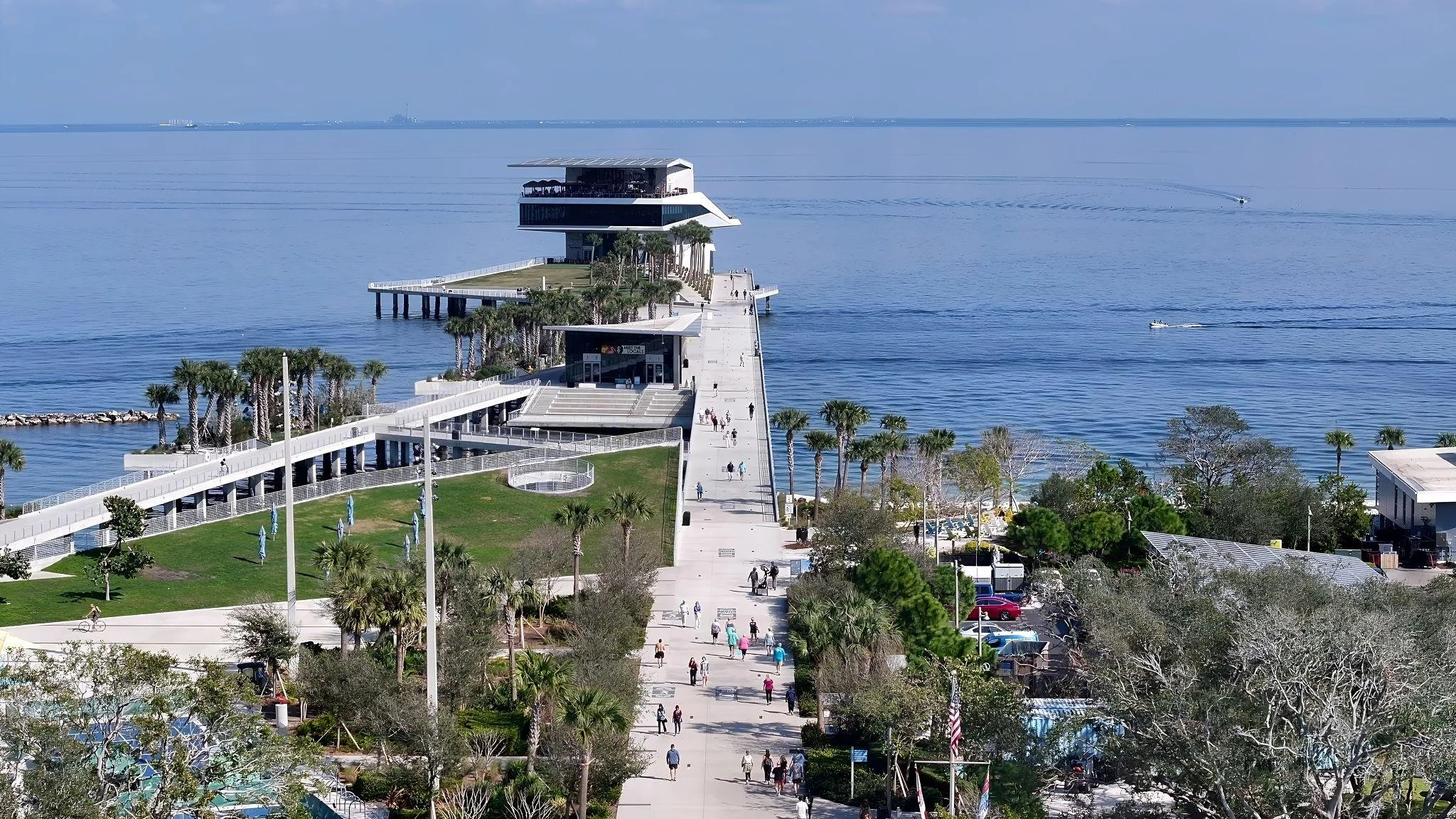 A pier extending into calm blue water, with a modern building at the end, lined with palm trees and a walkway with people walking, biking, and enjoying the area.