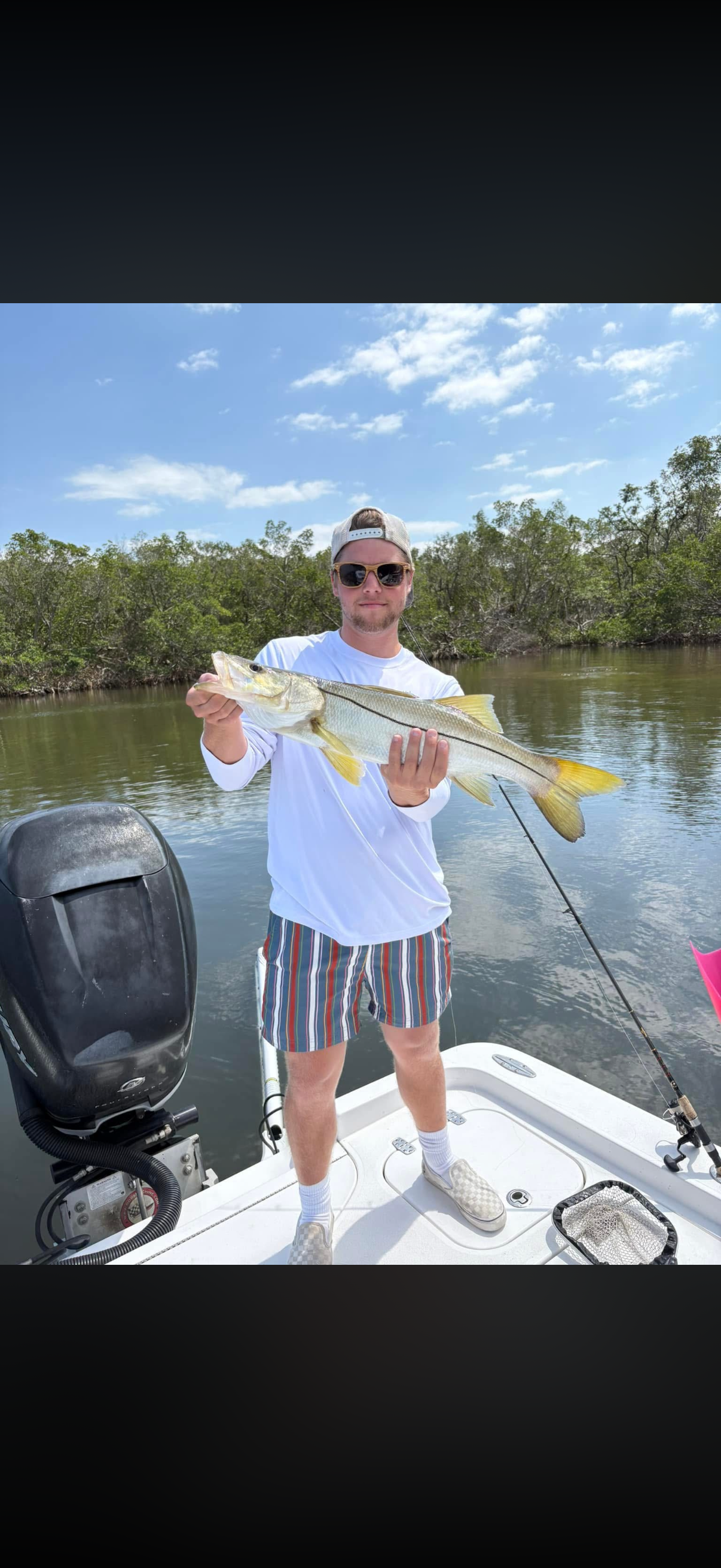Man in sunglasses wearing a white long-sleeved shirt, striped shorts, and a baseball cap on a boat holding a large fish with a yellow tail against a backdrop of water, trees, and a partly cloudy blue sky.