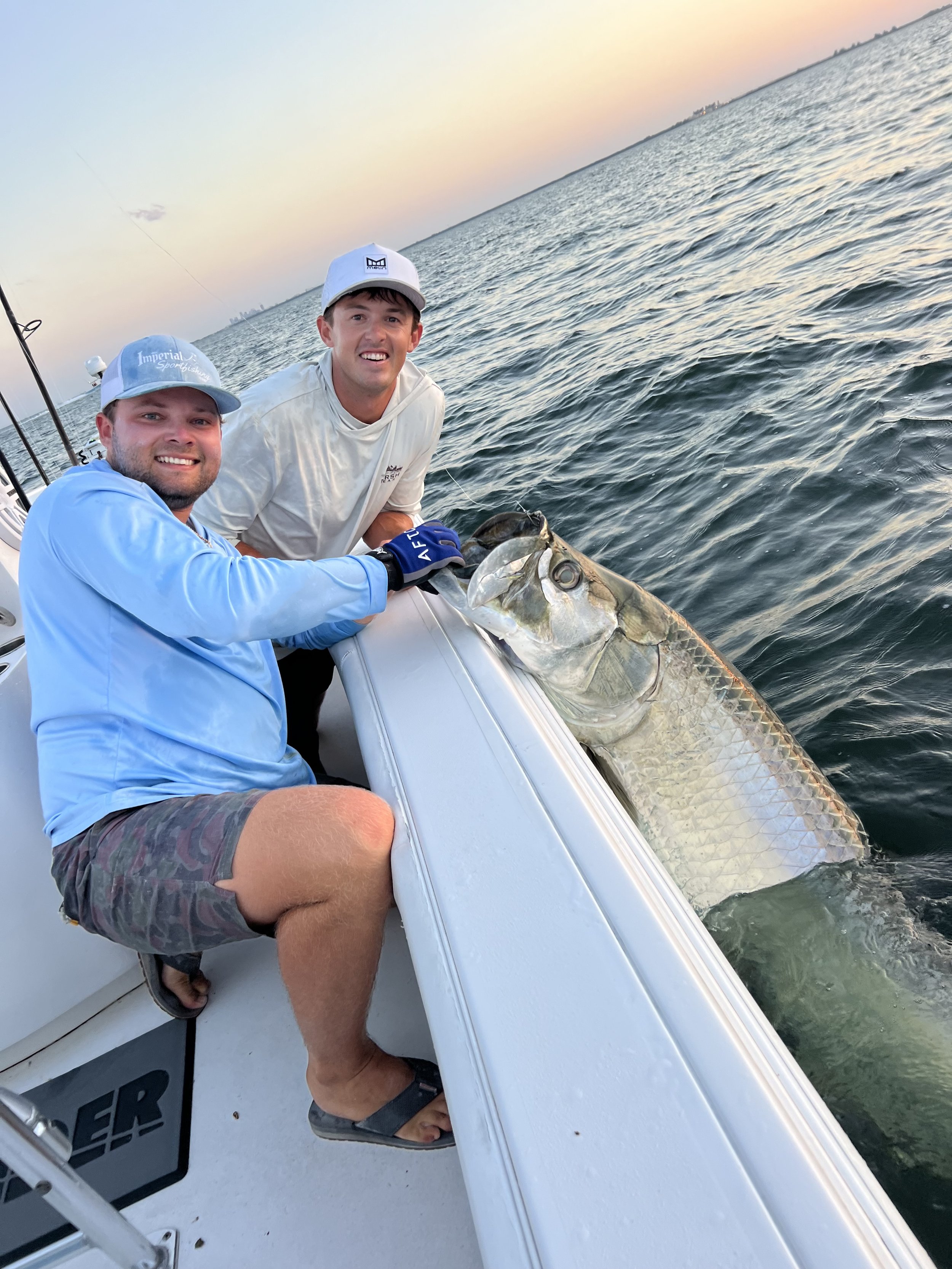 Two men on a boat holding a large fish they caught, smiling at the camera as the sun sets over the water.
