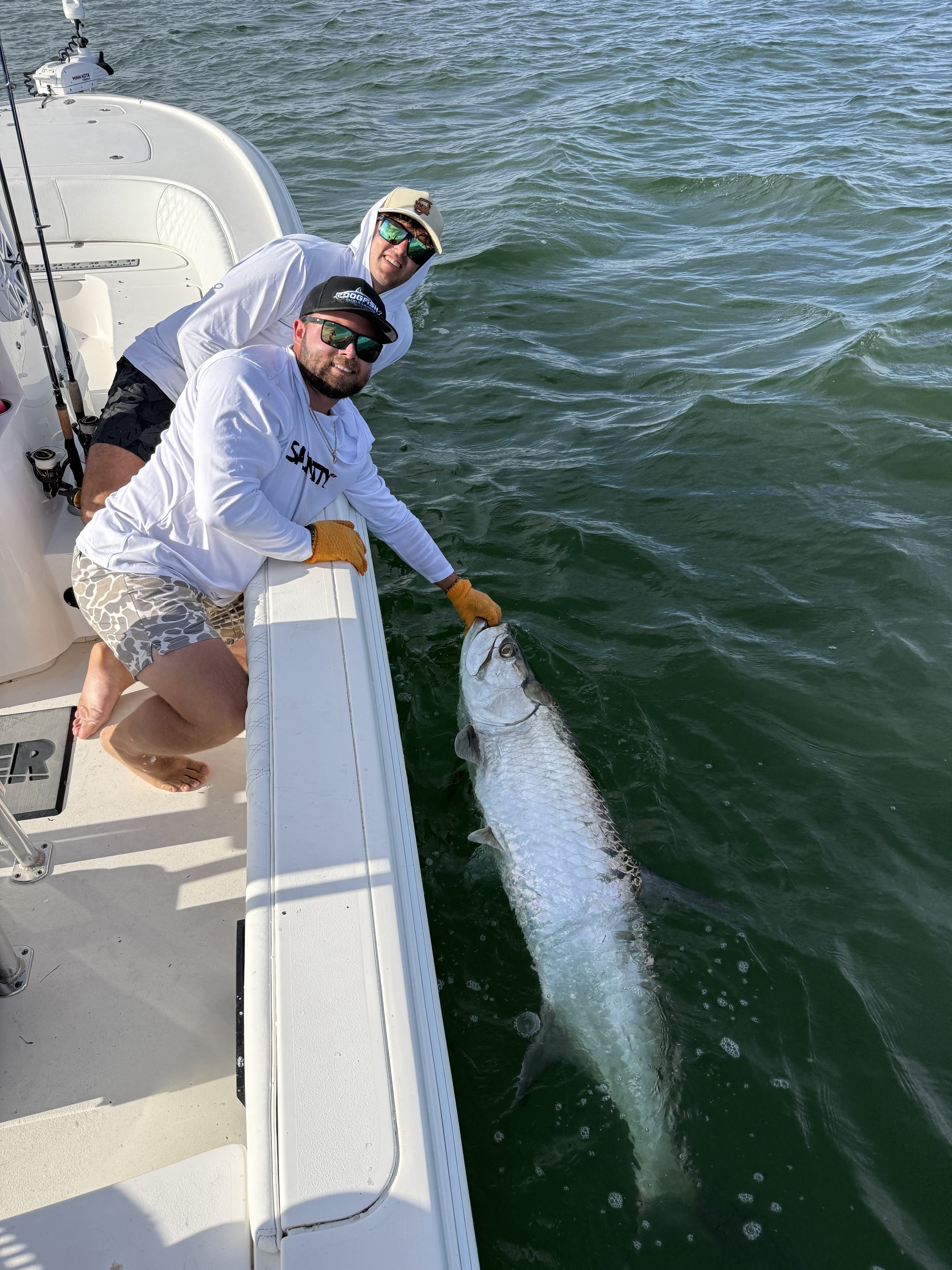 Two men fishing off the side of a boat with a large fish caught in the water.