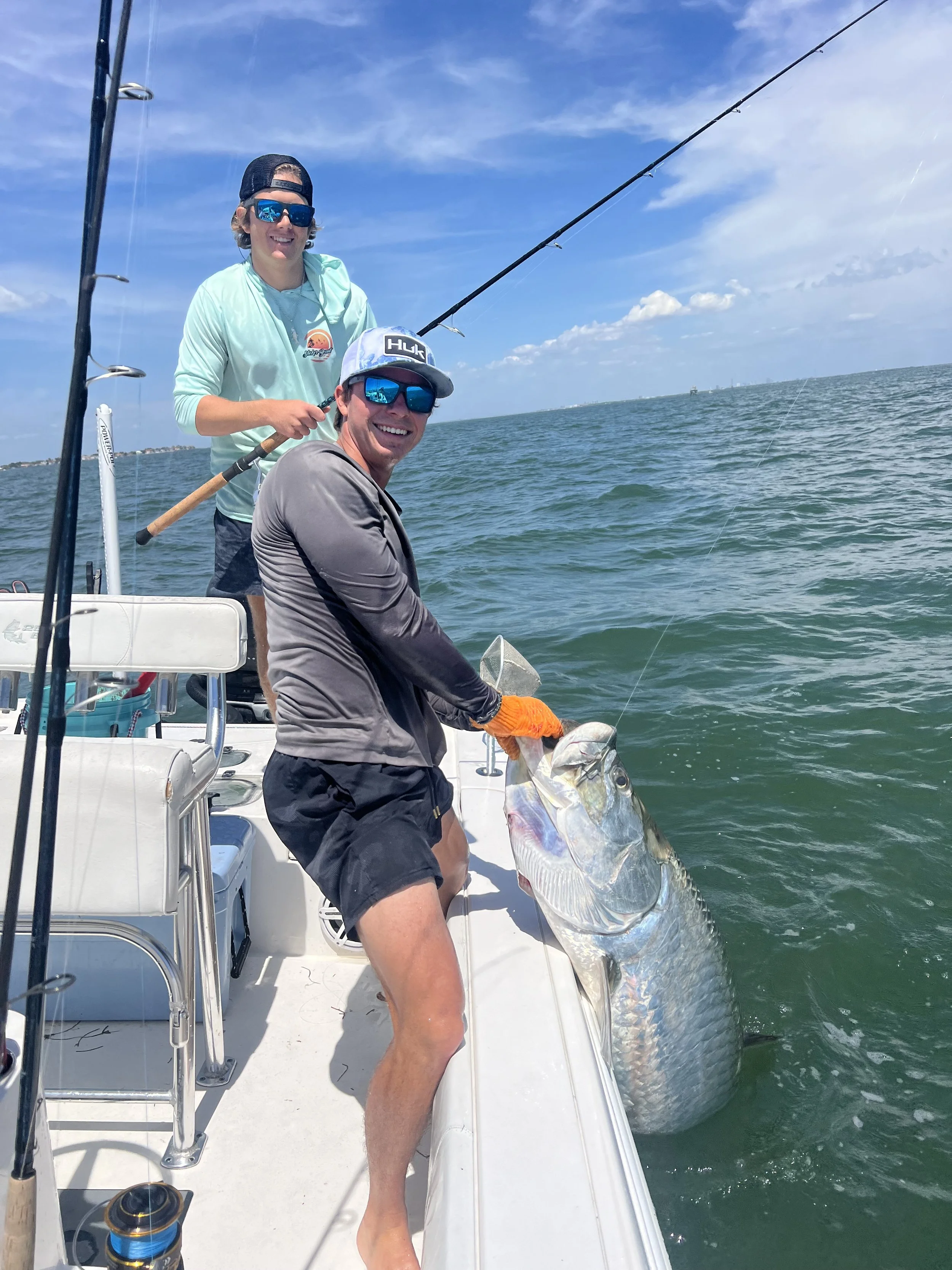 Two men on a boat fishing in the ocean with a large fish caught by one of them, smiling and wearing sunglasses under a partly cloudy sky.