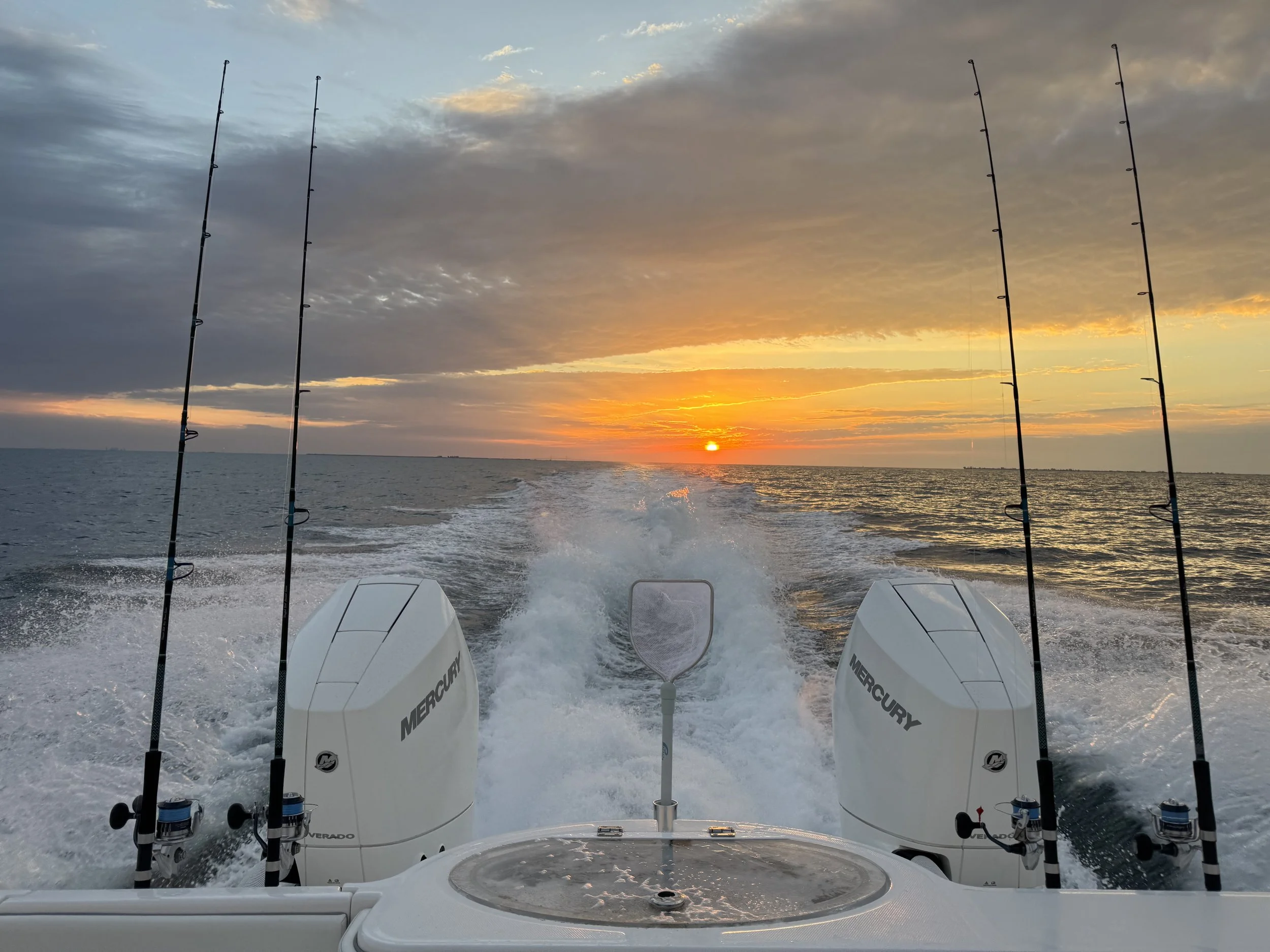 View from a boat with four fishing poles and two Mercury outboard motors at sunset. The ocean is calm with gentle waves.