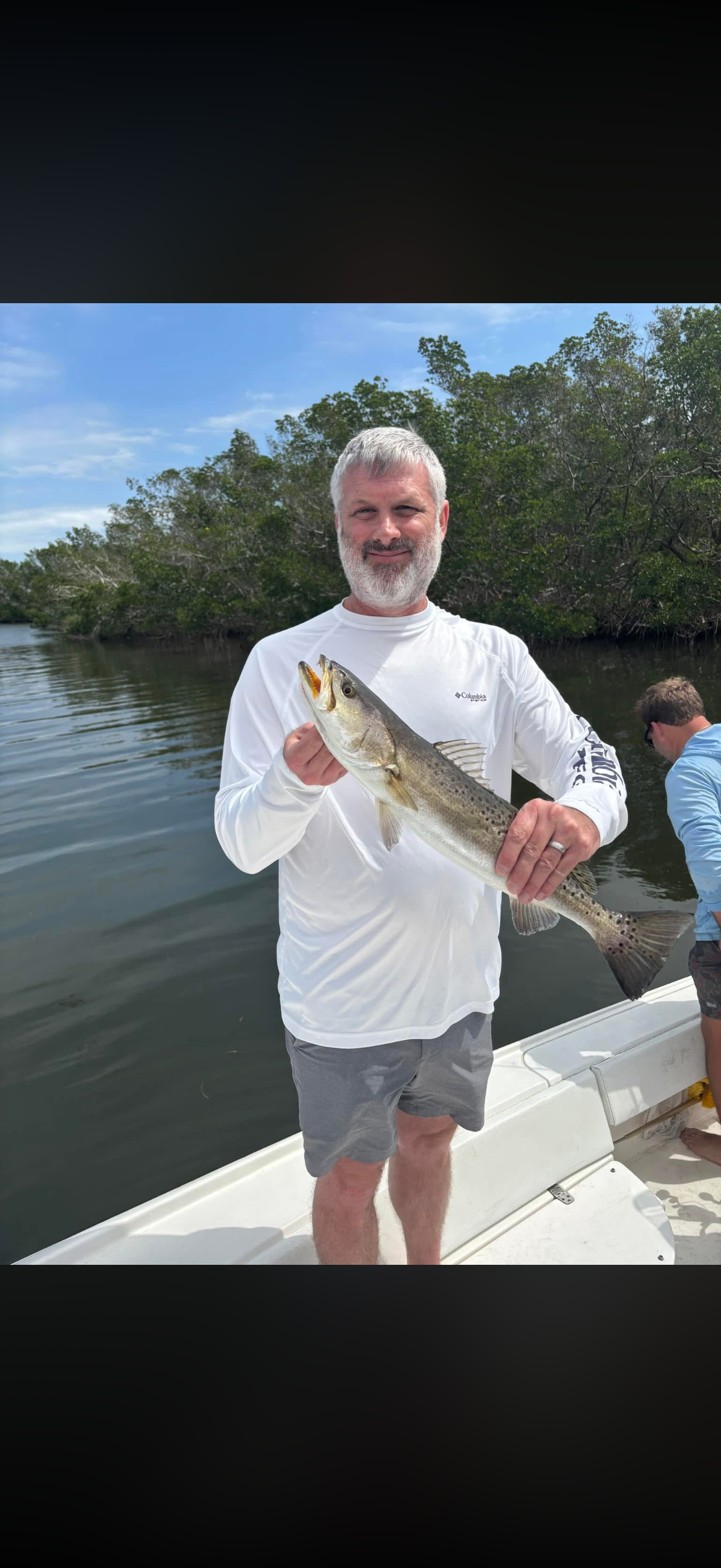 Man with gray hair and beard holding a large fish on a boat, with water and trees in the background.