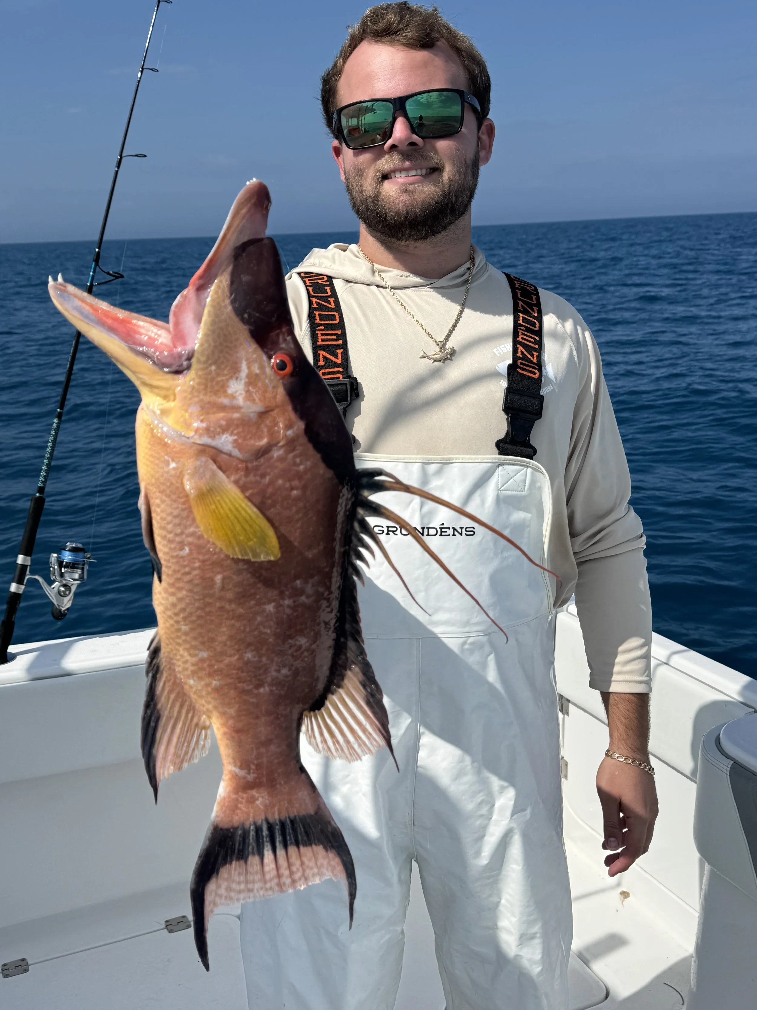 A man on a boat holding a large fish he caught, wearing sunglasses, a light-colored hoodie, and overalls, with fishing rods in the background and the ocean behind him.
