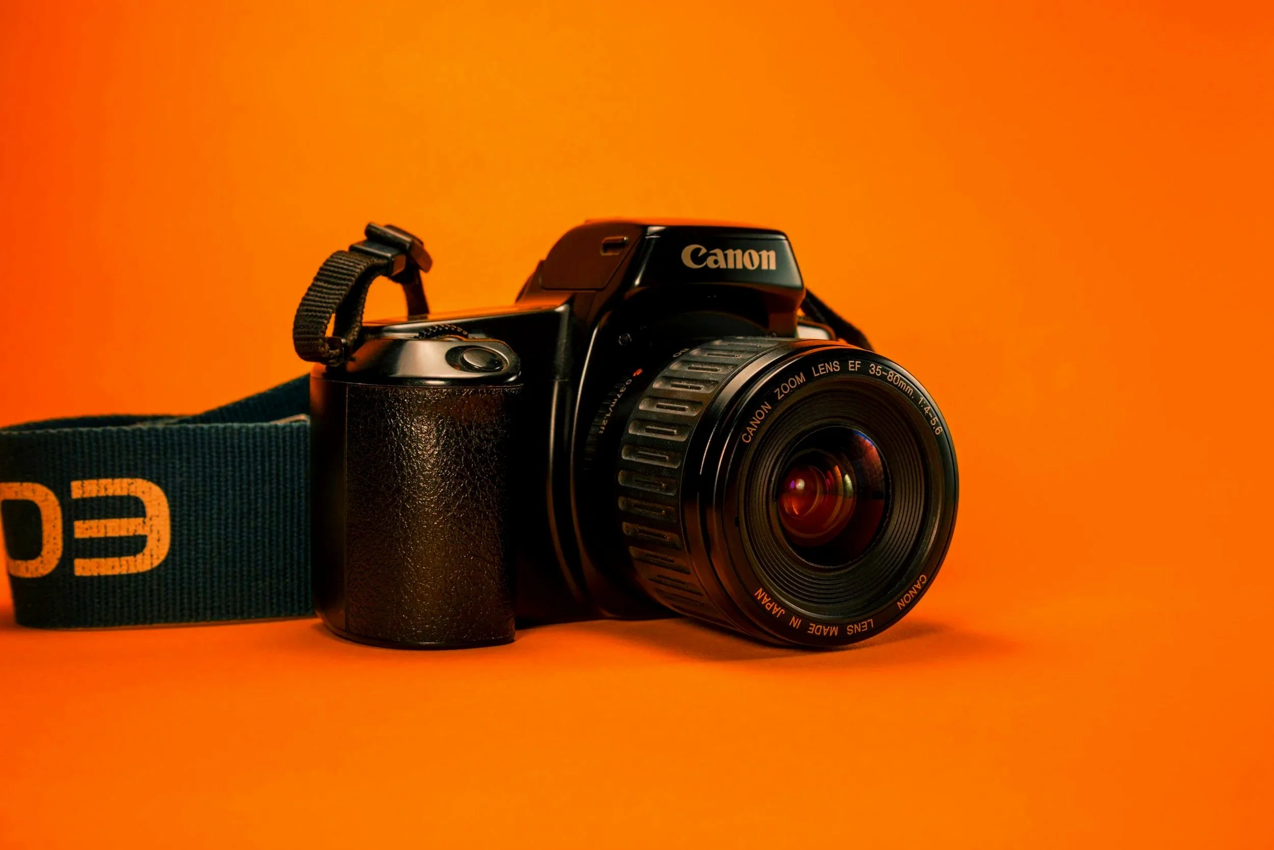 A black Canon camera with a zoom lens against an orange background.