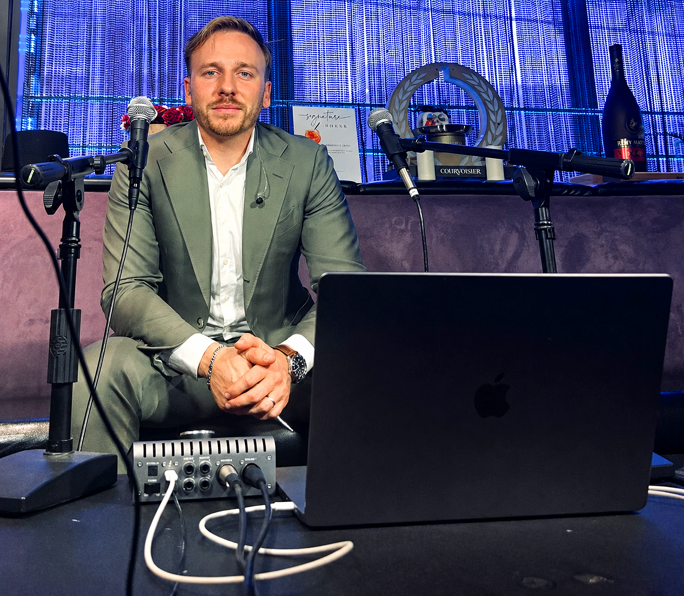 A man in a gray suit sitting at a table with audio equipment, microphone, and a laptop in front of him during a podcast or interview, with a blue-lit decorative background.
