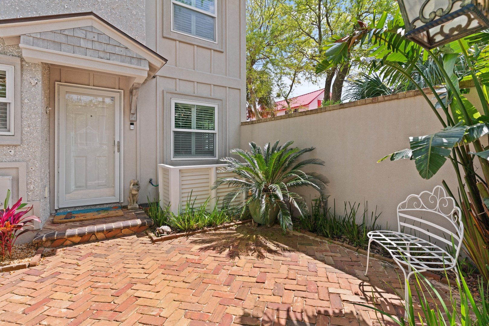 A small backyard patio with a brick floor, plants, and a white metal bench, adjacent to a beige house with a white door and windows, enclosed by a beige wall and trees in the background.