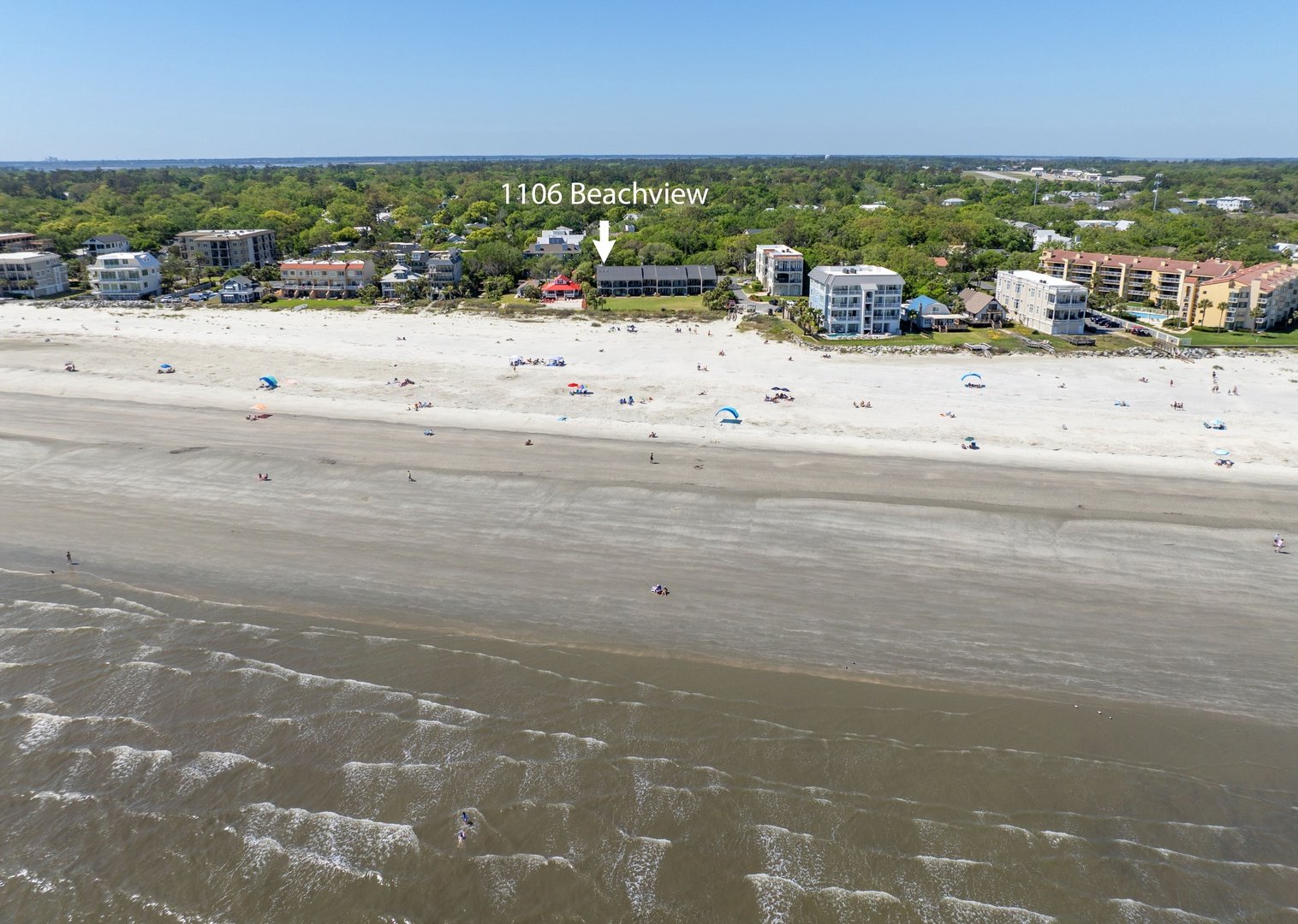 Aerial view of a beach with umbrellas, people, and a row of beachfront townhomes behind the sand dunes, labeled '1106 Beachview'