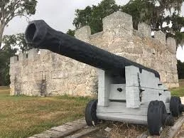 A large black cannon mounted on a gray wooden carriage with wheels, positioned outdoors near an old stone structure.
