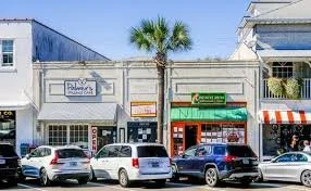A row of shops with cars parked in front, including a business named Palmers, a green storefront, and a building with a red and white striped awning, under a palm tree and blue sky.