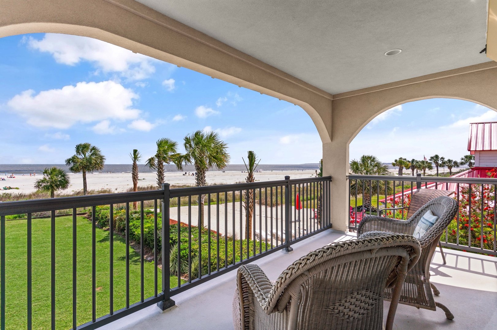 Balcony with wicker chairs overlooking a beach with palm trees and a clear blue sky.