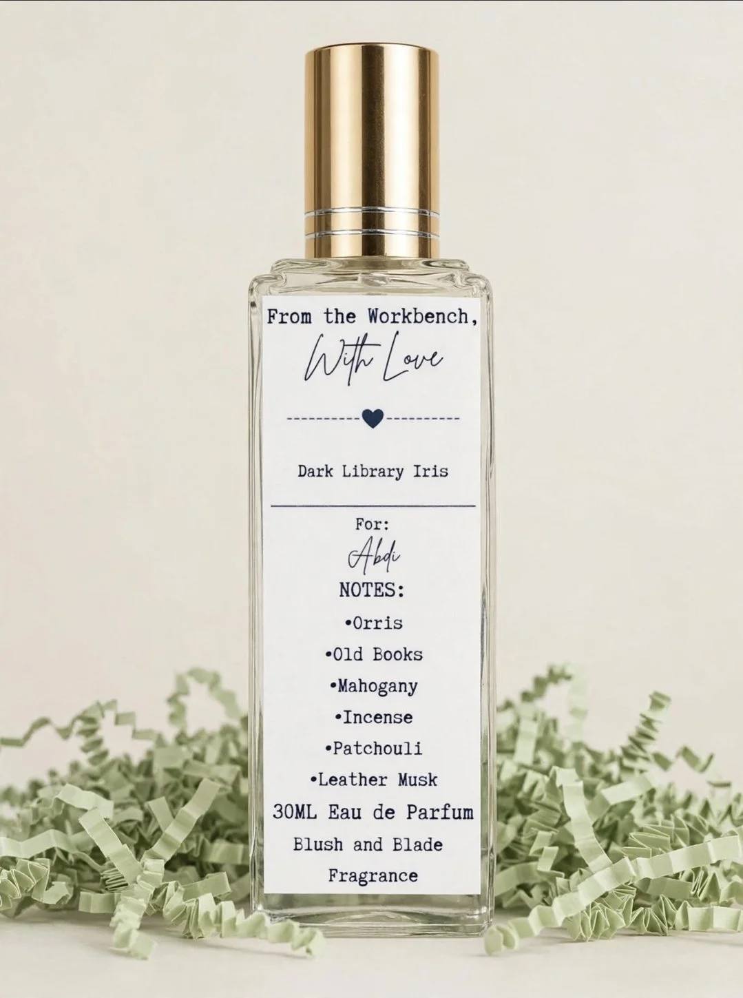 A clear perfume bottle with a gold cap and a label that reads 'From the Workbench, With Love' and lists fragrance notes including Orris, Old Books, Mahogany, Incense, Patchouli, Leather Musk, and Eau de Parfum for Abdi. The bottle is placed on shredded green paper.
