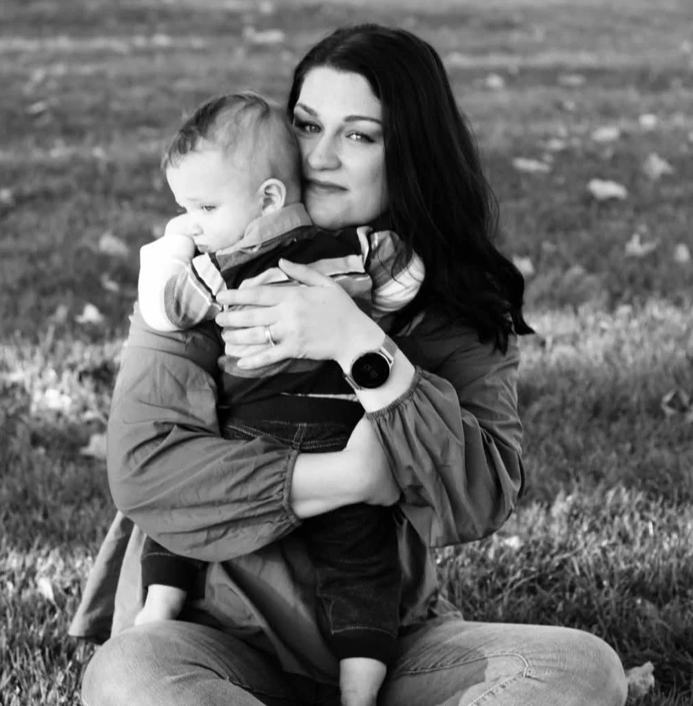 A woman with dark hair sitting on grass, holding a young child in a hug. The woman is wearing a jacket and a watch, the child is wearing a shirt and pants. They are outdoors with fallen leaves on the ground.