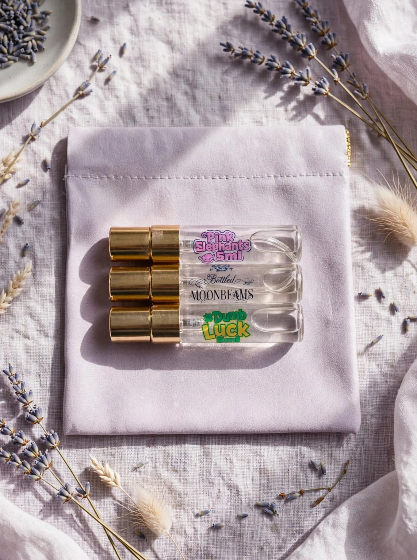 A flat lay of four small perfume bottles with gold caps labeled "Pink Elephants", "Bottled Moonbeams", and "Dumb Luck", placed on a white cloth napkin with dried lavender and other flowers surrounding it.