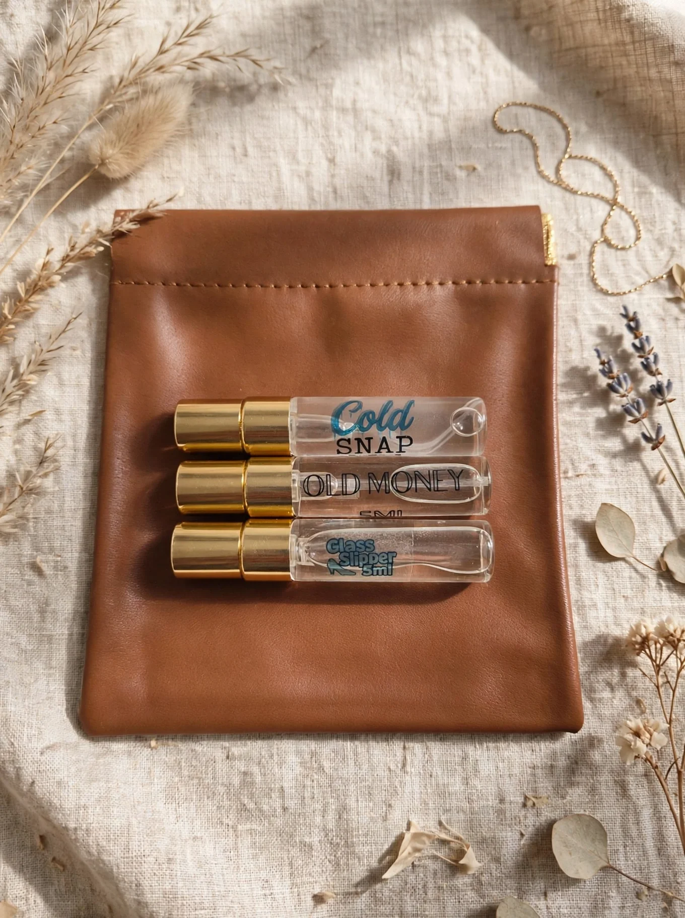 Clear bottles with gold spray caps and black text labels, placed on a brown leather pouch on a beige fabric surface, surrounded by dried flowers and leaves.