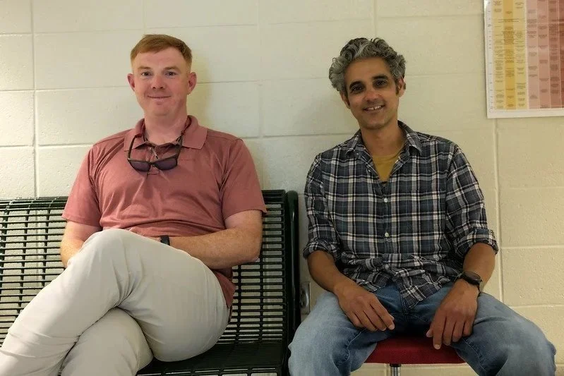 Two men sitting on a bench in a room with a chart on the wall behind them.
