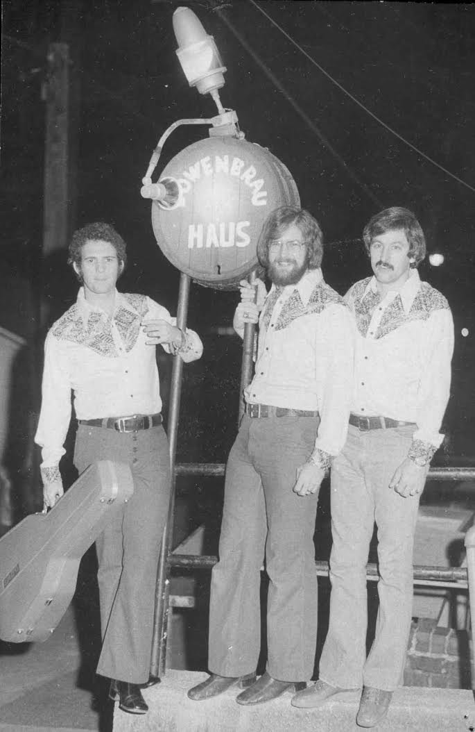 Black and white photo of three men standing outdoors at night, holding a sign that says 'Diener Bau Haus.' The men are wearing matching patterned shirts with wide collars and flared pants, indicating a 1970s fashion style. One man is holding a large pipe or pole, and another has a briefcase. A large beer barrel or drum with a tap is hanging above them, with a small lamp attached to it, shining light on the scene.