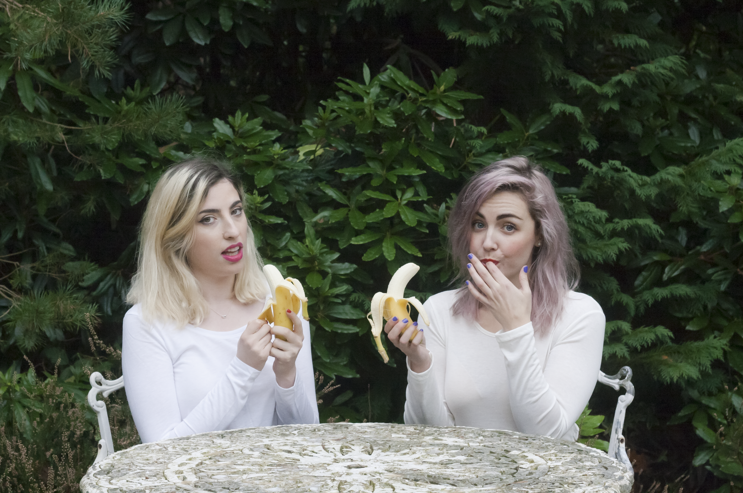 Two young women with light hair and wearing white shirts sitting at a stone table outdoors, eating bananas, with lush green plants and bushes behind them.