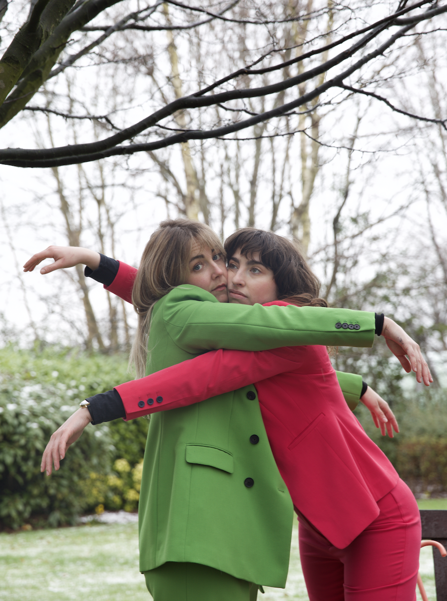 Two women wearing colorful suits, one in green and the other in red, embracing in an outdoor park with leafless trees in the background.