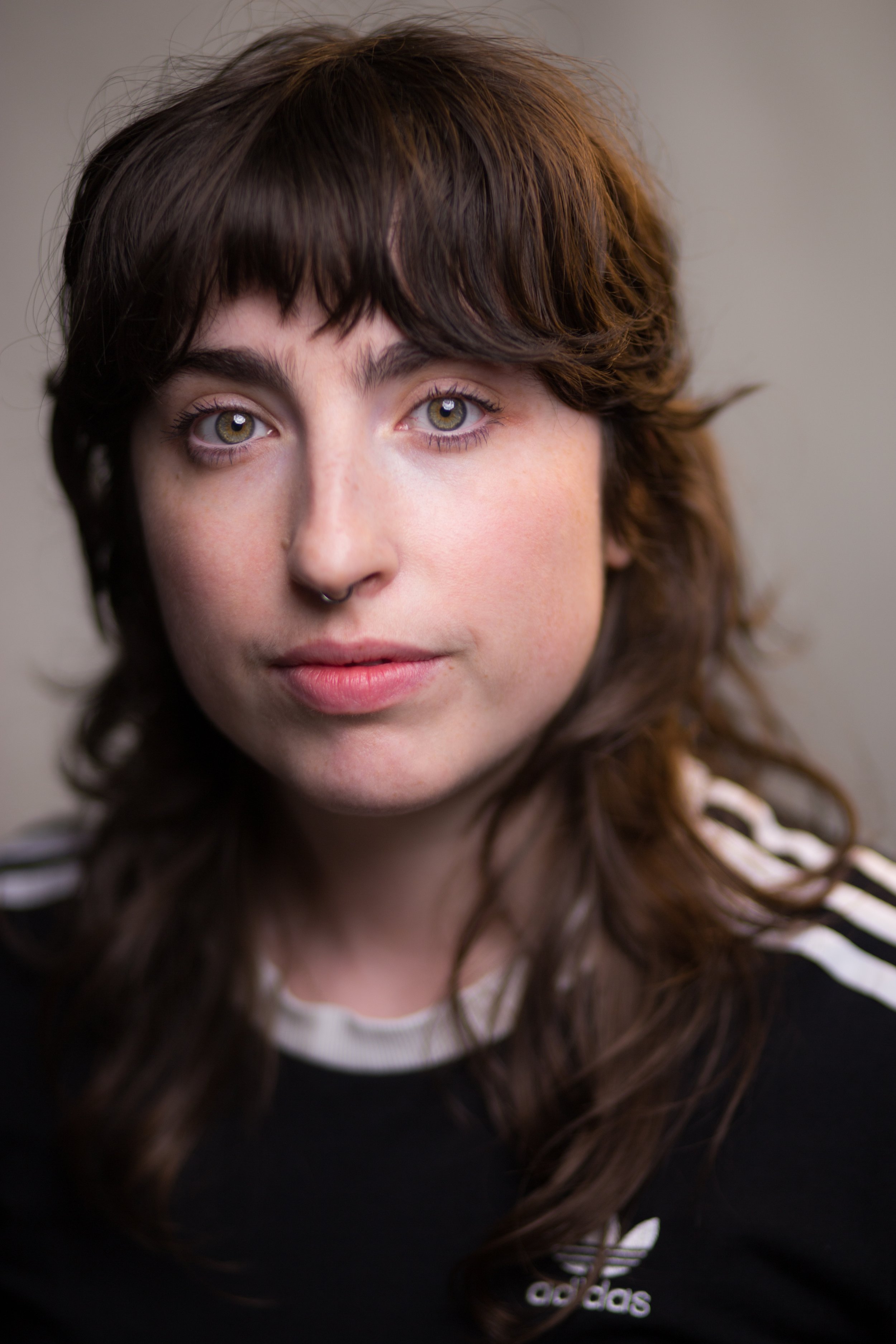 Close-up of a young woman with wavy brown hair and hazel eyes, wearing a black Adidas sweatshirt with white stripes and logo, against a plain background.