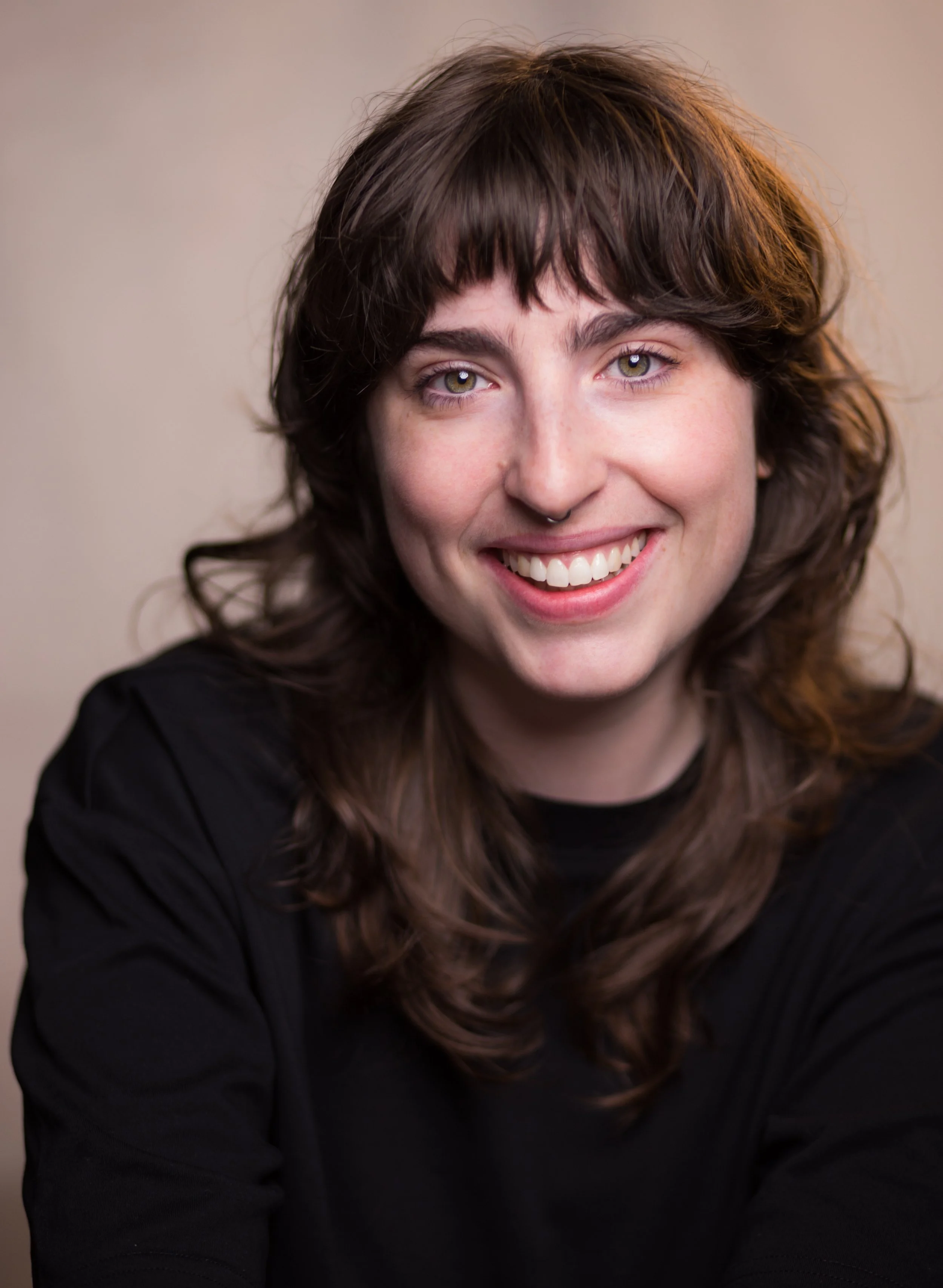 Close-up of a woman with long, wavy brown hair smiling, wearing a black top, against a neutral background.