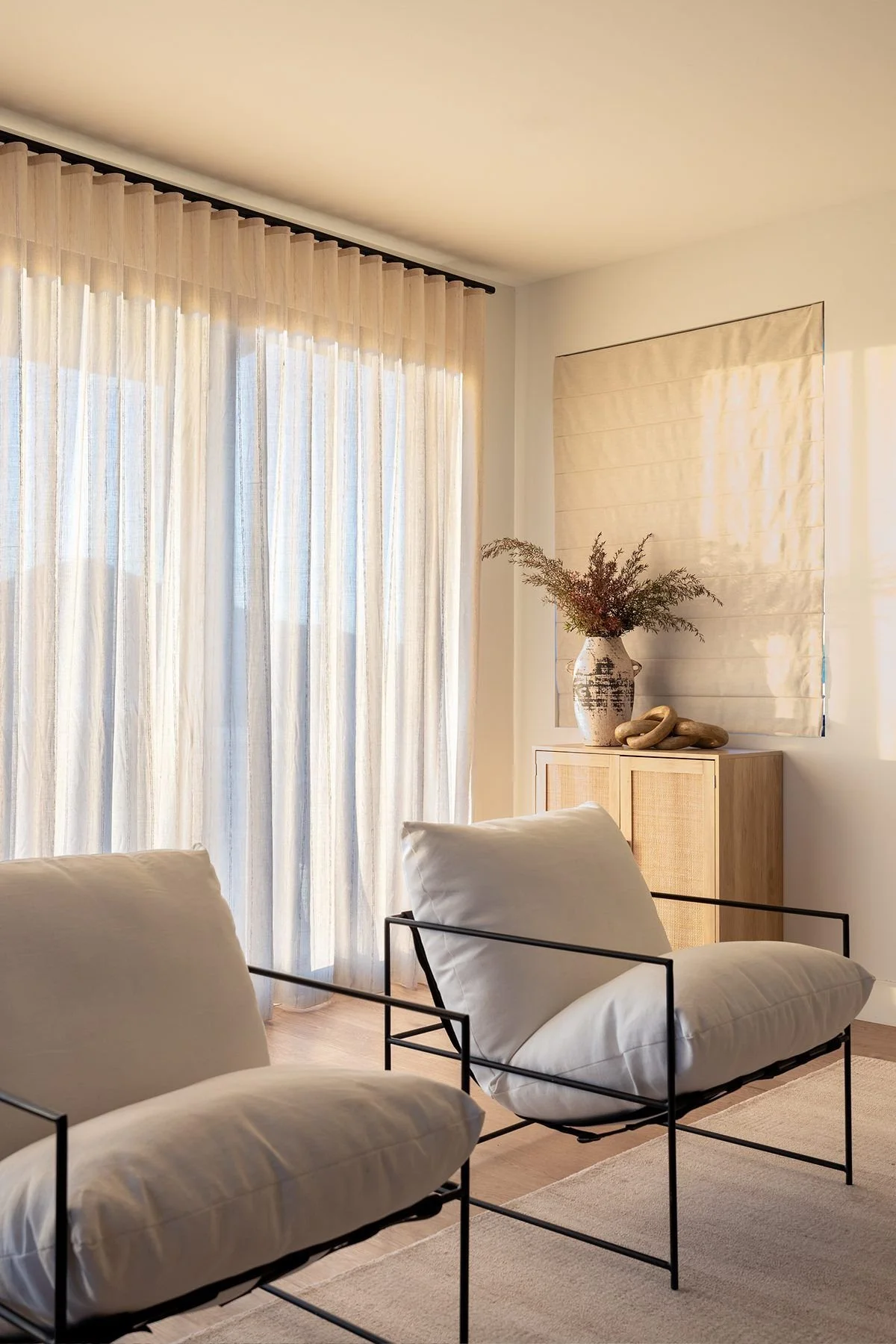 A room with sheer white curtains covering large windows, beige walls, and a light wooden cabinet topped with a decorative vase, rocks, and dried branches. Two white cushioned chairs with black metal frames are in the foreground, and a beige rug covers the wooden floor.