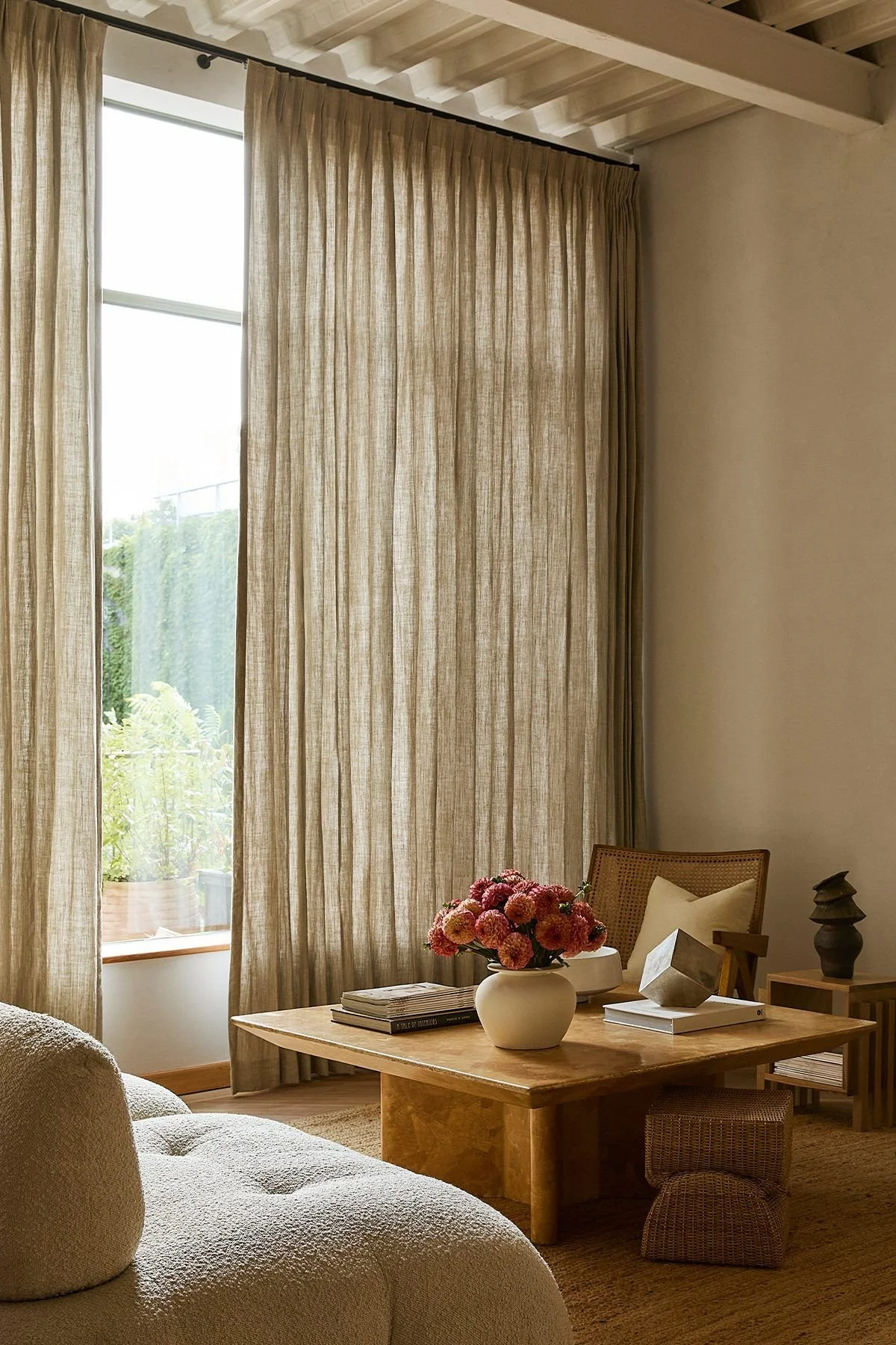 Living room with beige curtains covering a large window, a wooden coffee table with a white vase of pink flowers, a stack of magazines, and decorative objects, a beige sofa in the foreground, a wooden side table with more decor, and a chair with a cushion.