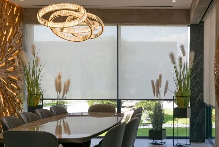 Modern conference room with a large table, stylish chairs, tall green plants in planters, large window with blinds, and elegant circular light fixtures overhead.