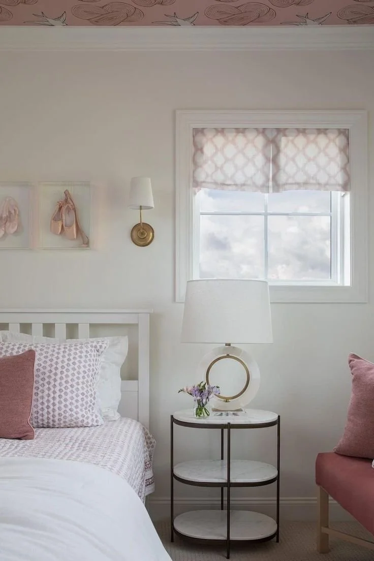 A cozy bedroom corner with a white bed, pink and white pillows, a nightstand with a white lamp, a small flower vase, and pink upholstered chair, a window with a patterned shade, and soft pastel decor.