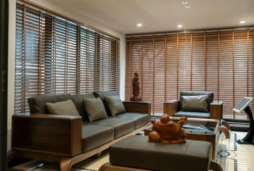 Living room with wooden blinds, a gray sofa with beige pillows, a matching armchair, a wooden side table with a tall carved wooden sculpture, a glass-topped wooden coffee table, a decorative wooden sculpture on an ottoman, and a digital tablet resting on a stand.